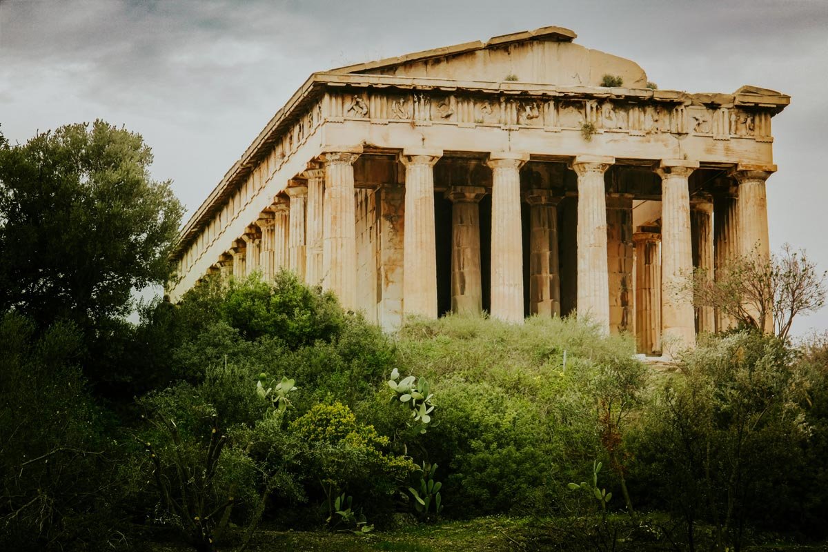 Greek Doric temple emerging from dense green trees on a hill, lit by soft afternoon light beneath a cloudy sky.