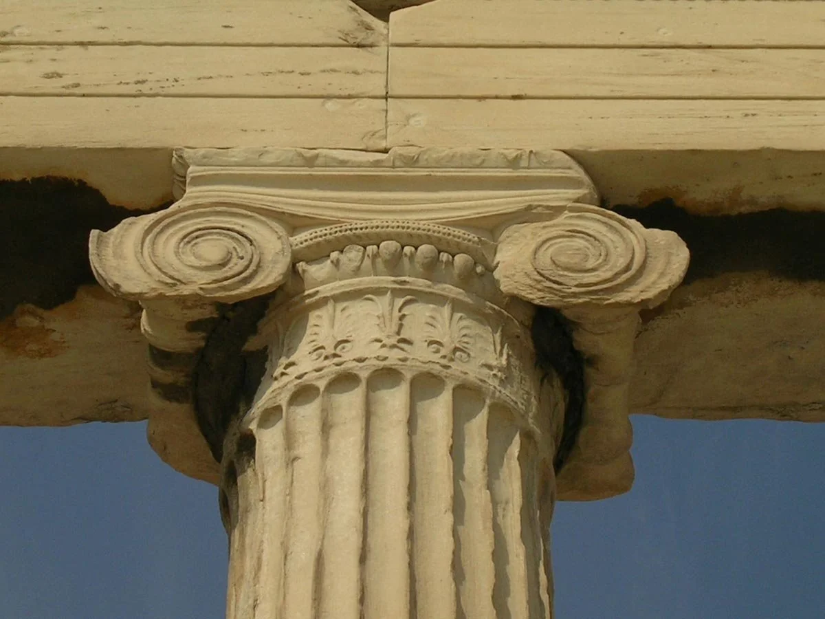 Close-up of an Ionic column capital, with volute scrolls, egg-and-dart molding and carved leaves beneath a weathered architrave.