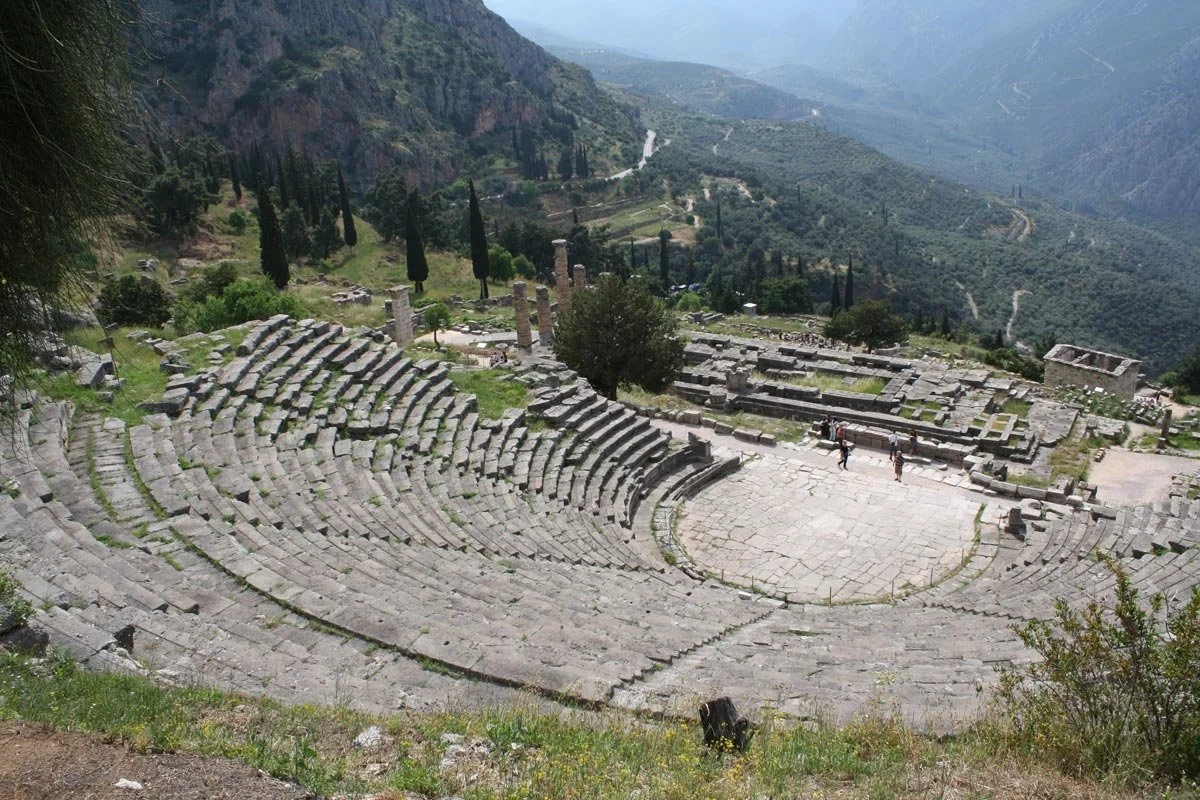 Ancient stone theater at Delphi seen from above, semicircular seating cut into the hillside overlooking sanctuary ruins and valley.