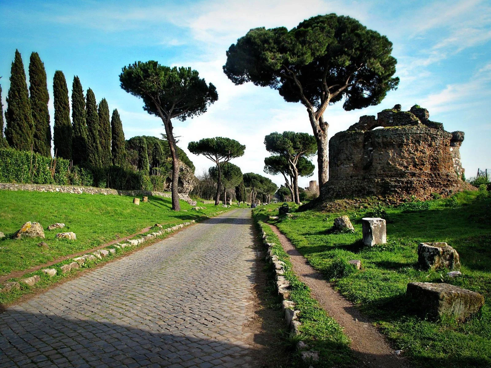 Straight paved stretch of the Via Appia Antica lined with grass, trees, and ancient remains.