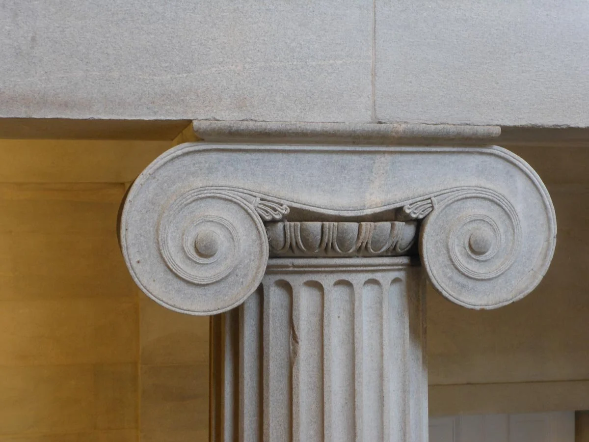 Close view of a grey Ionic column capital, with crisp spiral volutes and fluted shaft inside a classical interior.