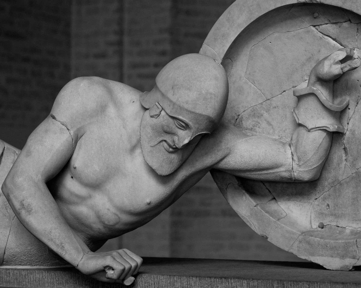 Fallen warrior from the Temple of Aphaia, propped on one arm with shield, muscular body tense in near collapse.