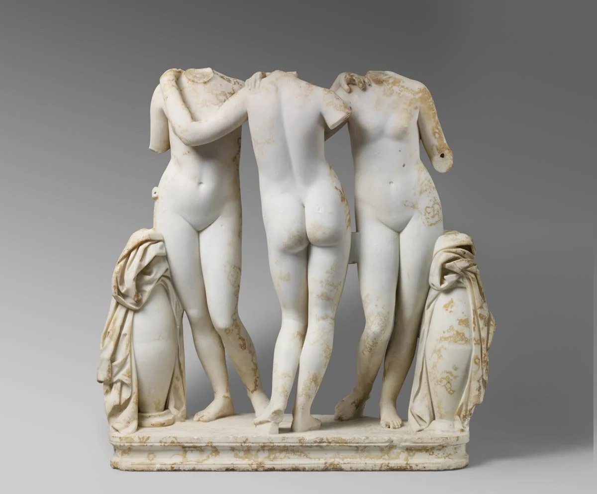 Marble group of three nude female figures standing close together with their arms linked across shoulders.