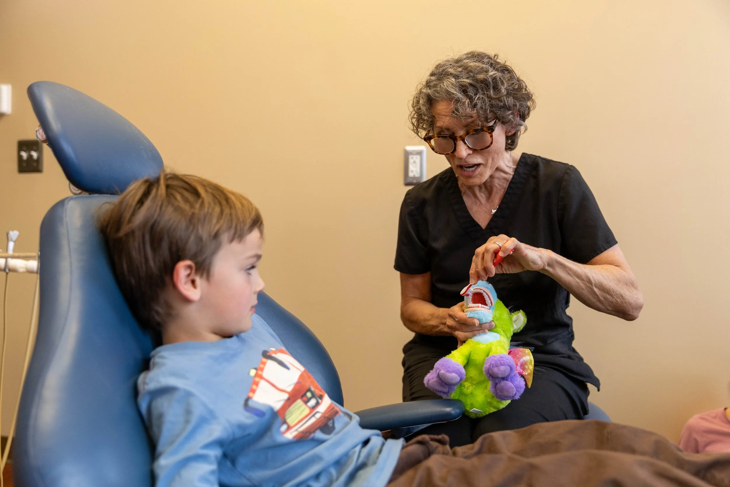 A young boy sitting in a hospital chair with medical equipment, and an older woman showing him a soft plush dinosaur toy, in a room with beige walls.