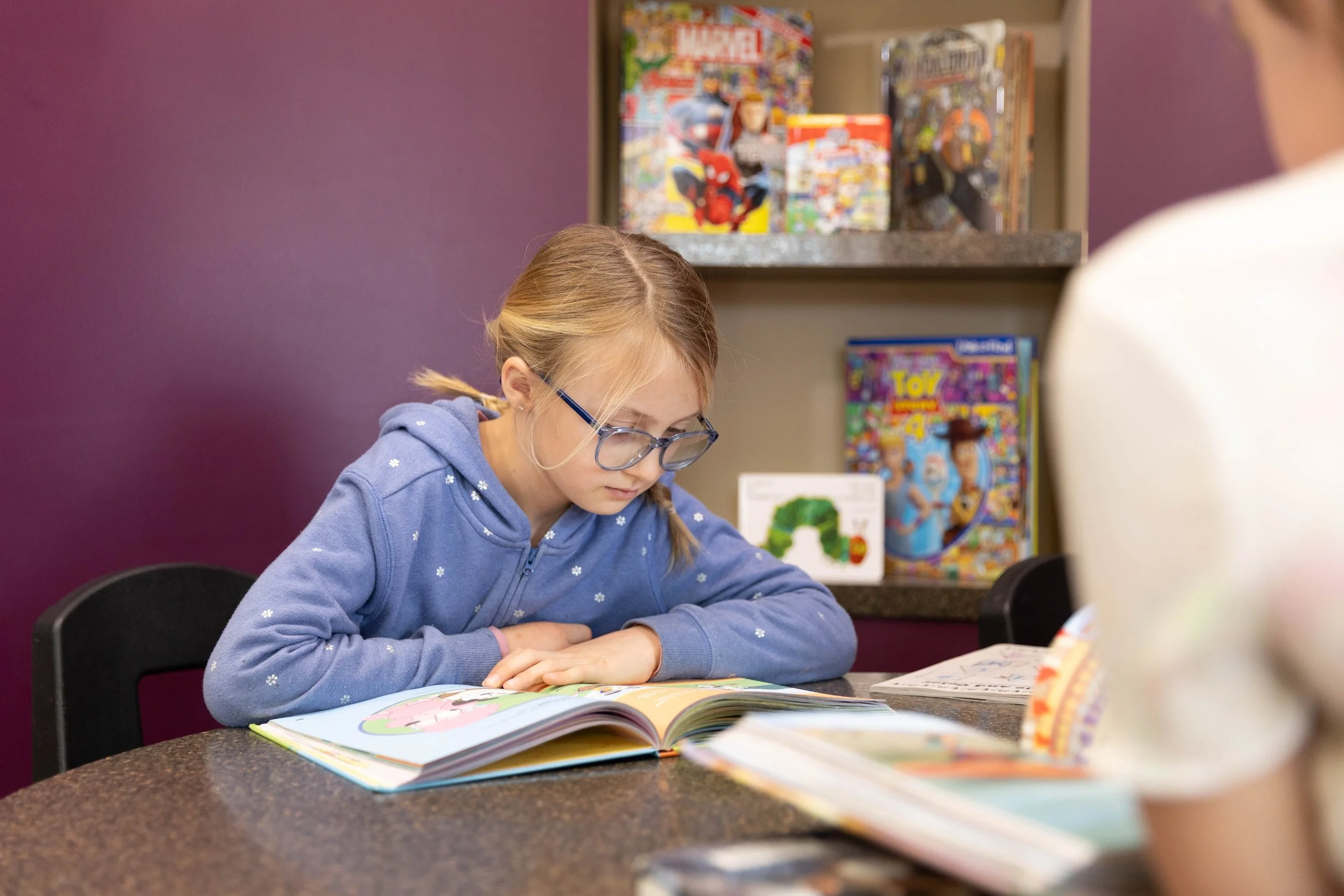 A young girl with glasses and blonde hair tied in pigtails, wearing a blue hoodie, is sitting at a table reading a colorful children's book. There are other books on the table and shelves in the background display comic books and a children's magazin