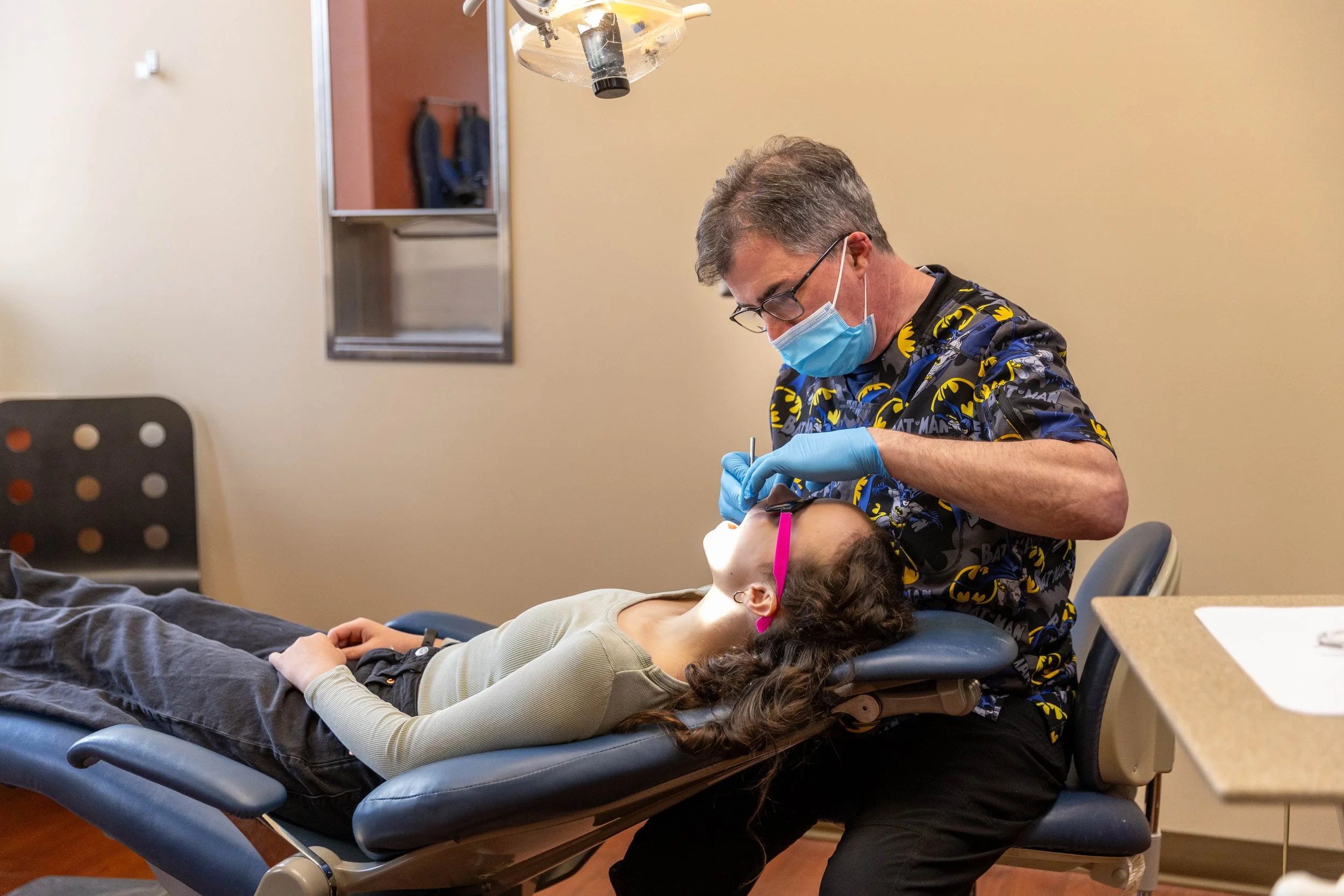 A dentist in a Batman shirt, wearing a face mask and gloves, performs a dental examination on a young girl with curly hair and pink glasses who is lying back in a dental chair.
