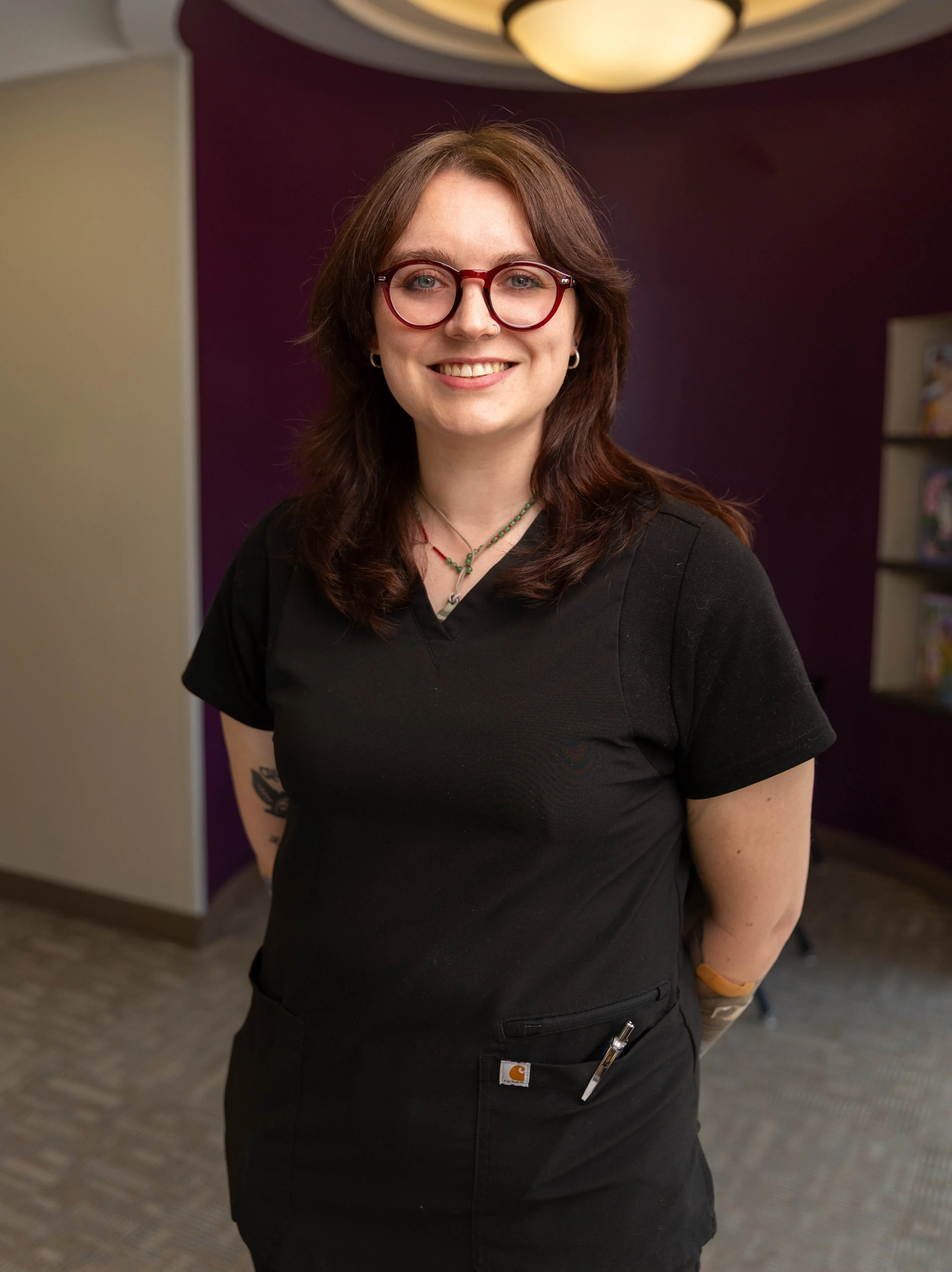 A woman with shoulder-length brown hair, glasses, and wearing a black t-shirt, smiling indoors with a purple wall in the background.