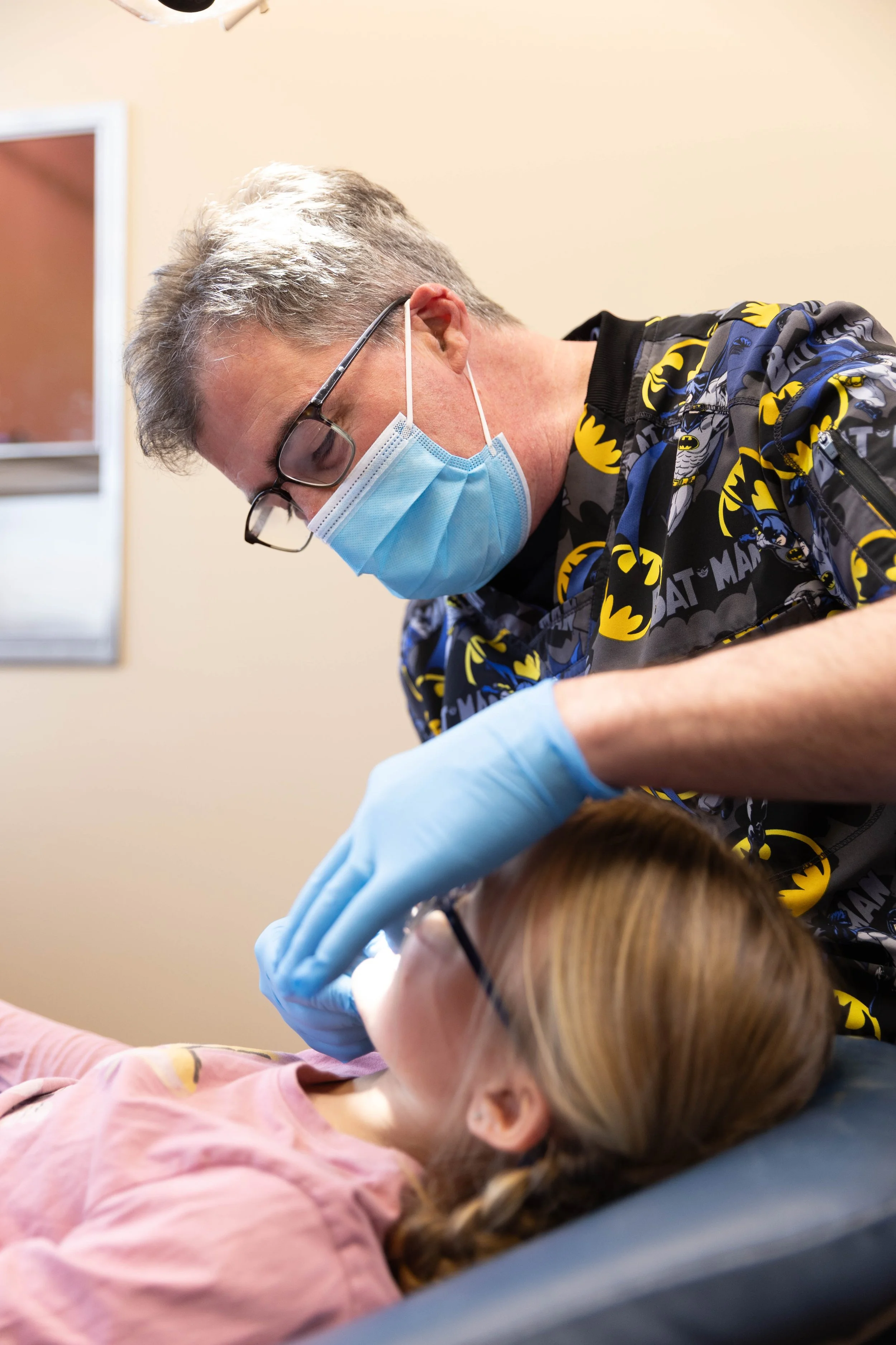 A dentist or oral health professional wearing a Batman-themed scrub top, glasses, a face mask, and gloves treating a female patient with red hair, glasses, and a pink shirt, in a dental clinic.