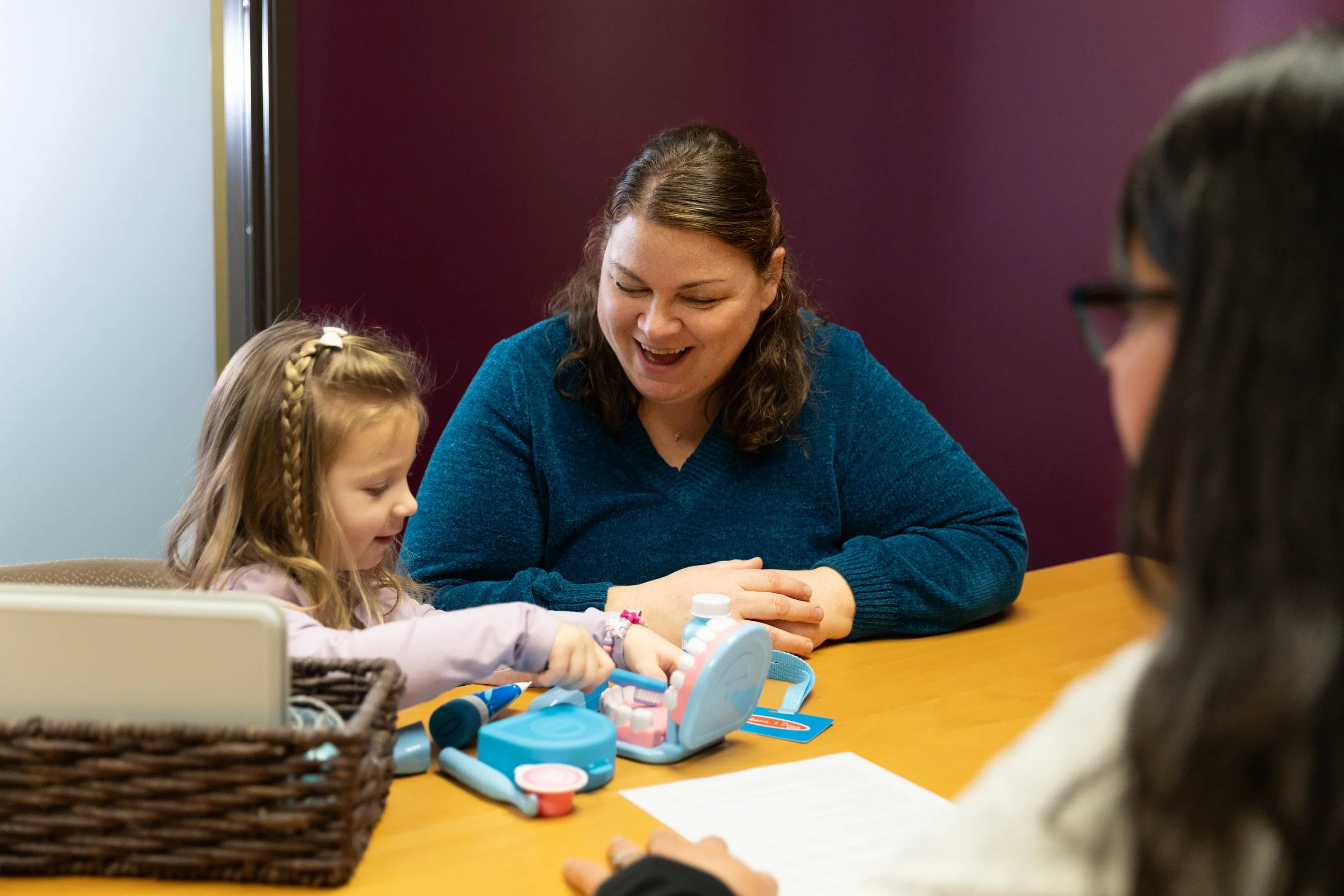 A woman and two girls sitting at a table, playing with miniature medical toys, with a basket and papers on the table.