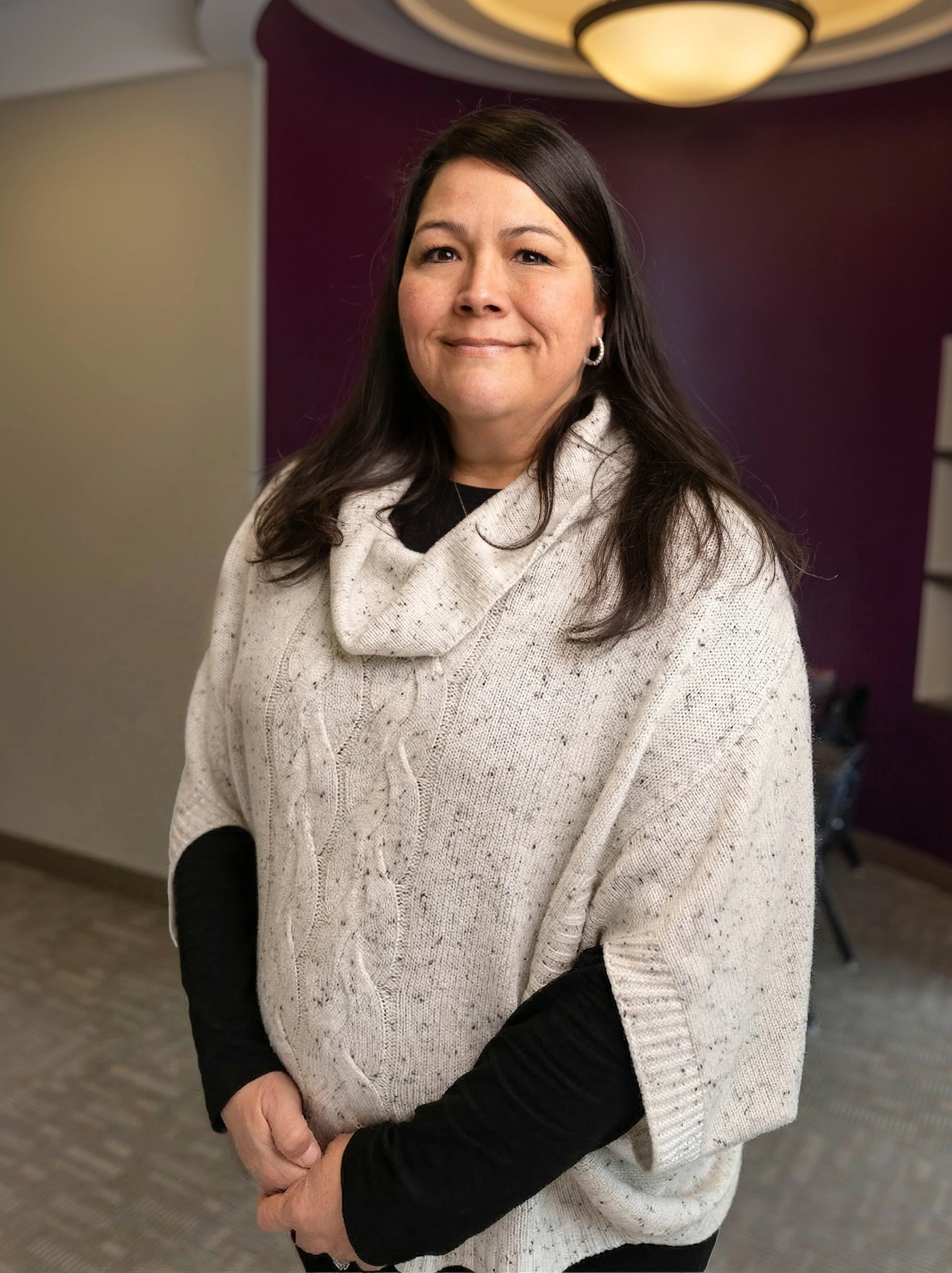 A woman with dark brown hair, wearing a cream-colored cable knit poncho over a black long-sleeve shirt, standing indoors with a dark purple wall and ceiling light in the background.