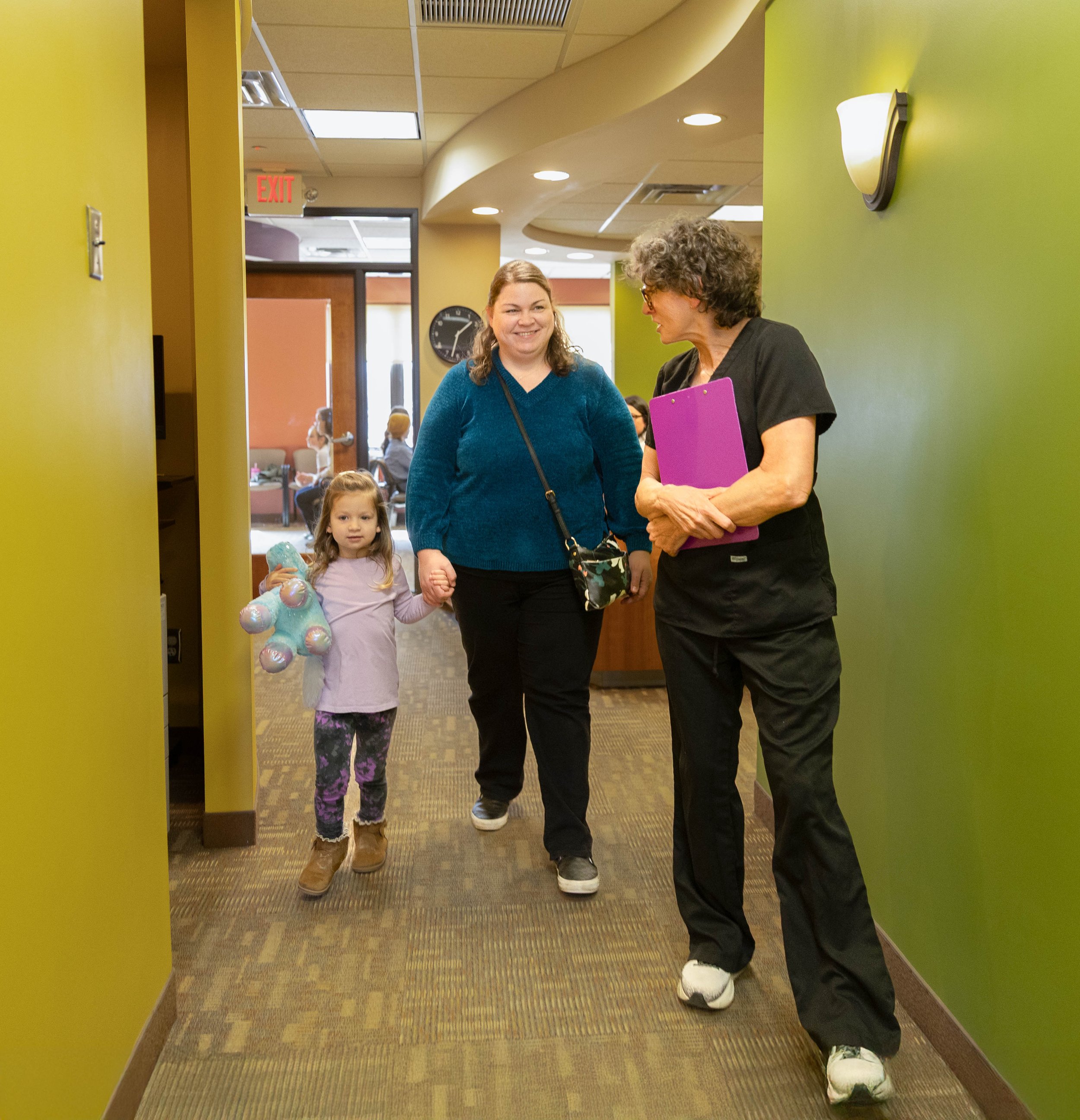 A woman in a blue sweater holding a child's hand, walking with a woman in black scrubs through a hallway in a medical or office building, with other people visible in the background.