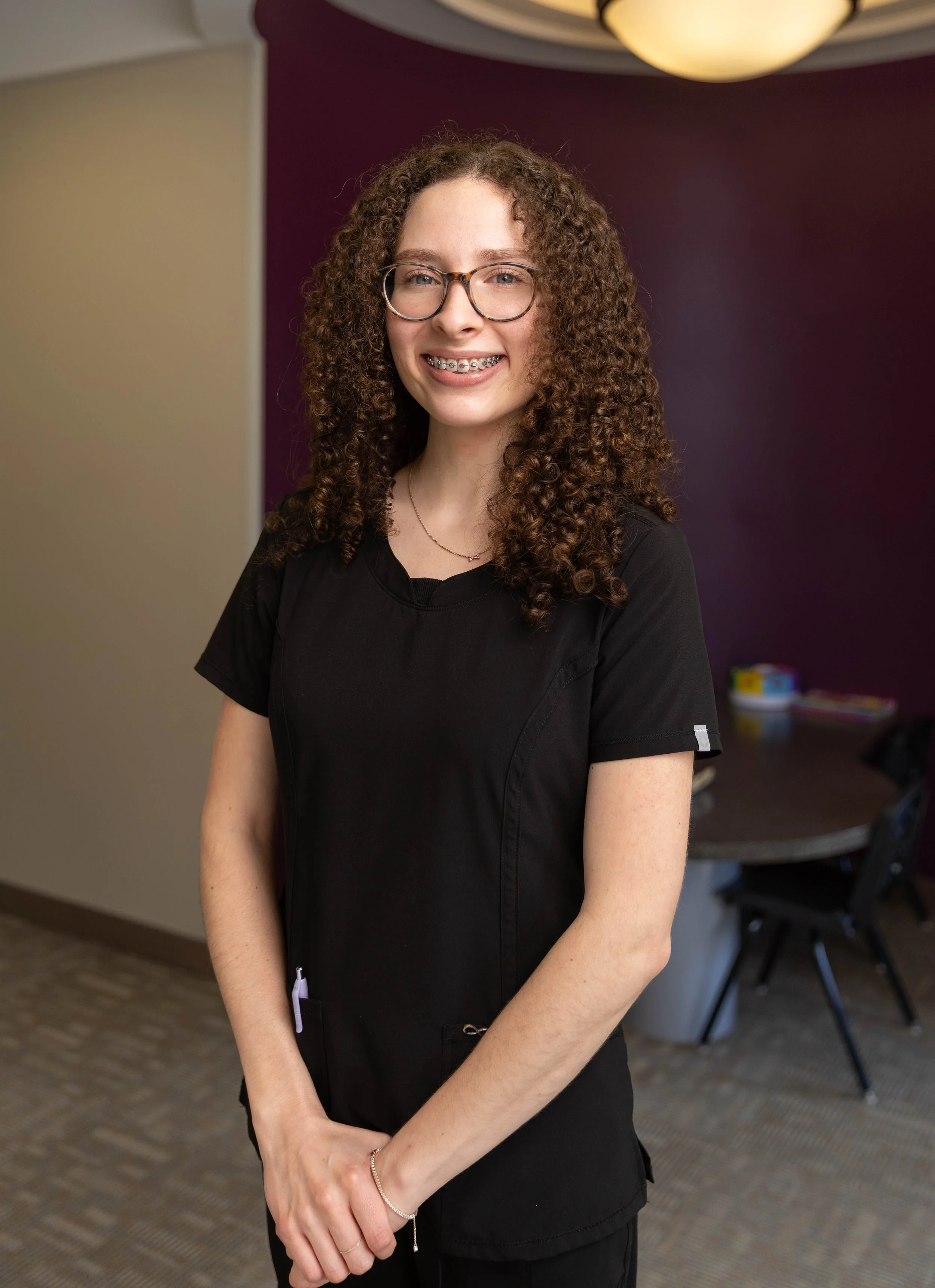 A young woman with curly brown hair and glasses smiling, wearing a black medical uniform, standing indoors with a purple and beige wall in the background.