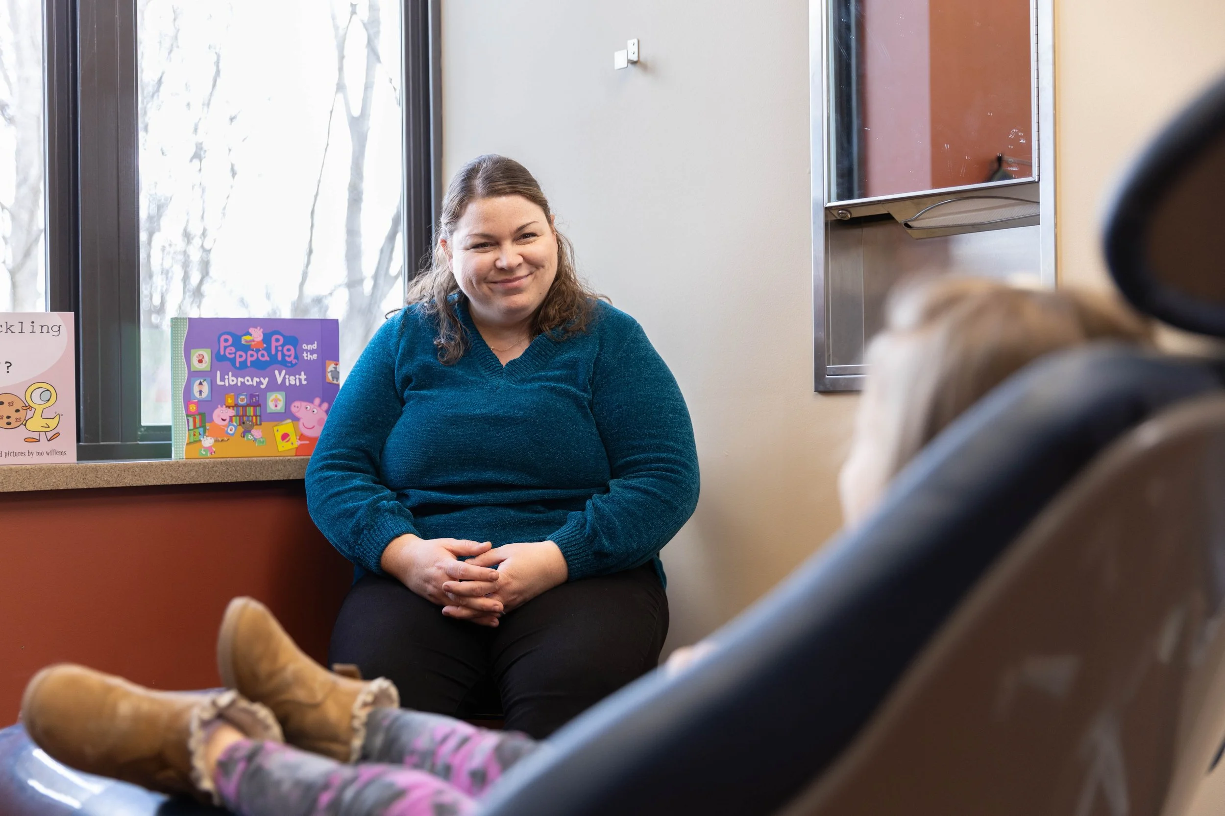 A woman sitting by a window, smiling at a child who is lying on a chair. The woman has brown hair and is wearing a teal sweater. The child is wearing tan boots and patterned leggings, with their head turned away from the camera. A children's book tit