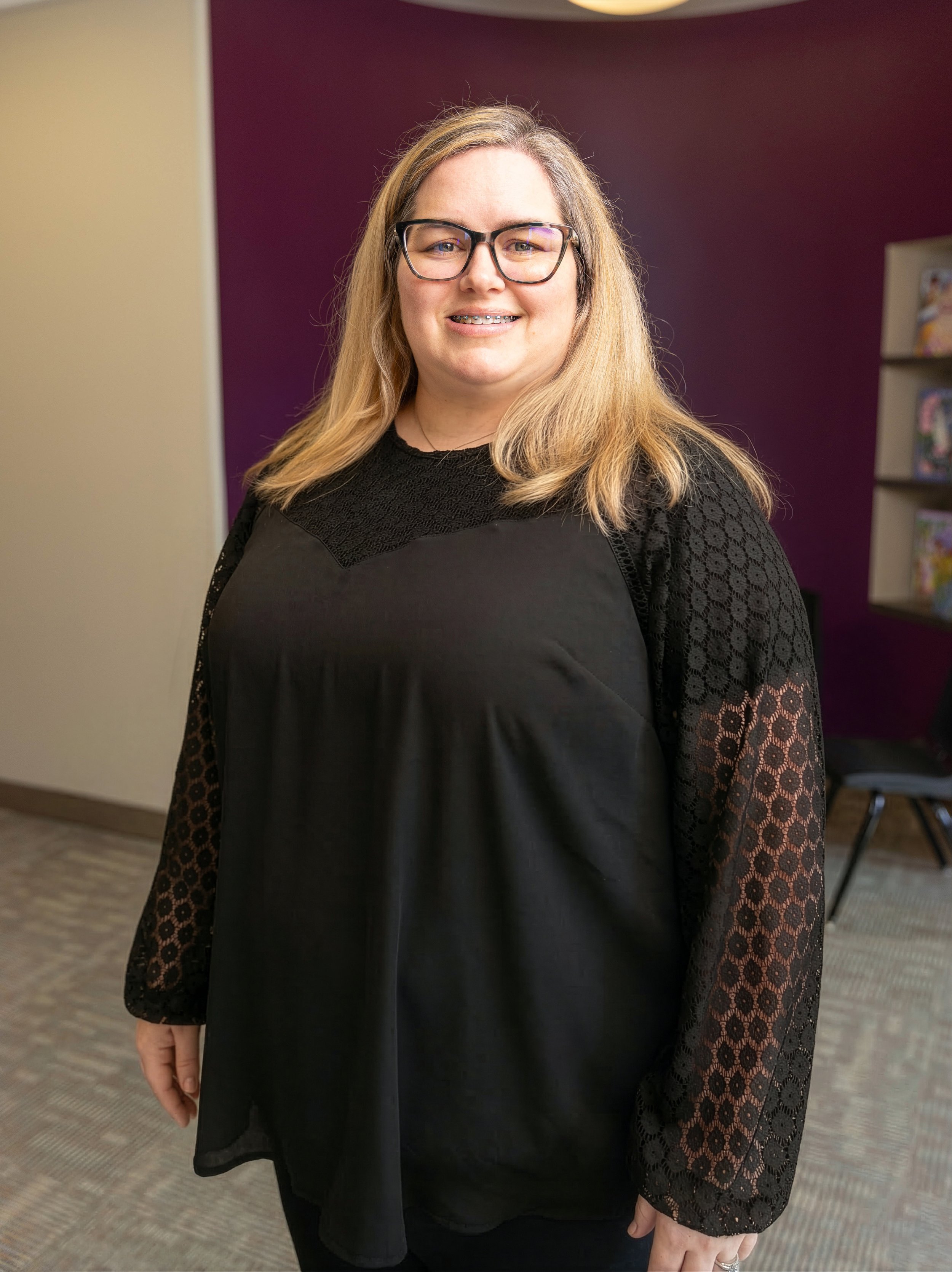 A smiling woman with glasses and long blonde hair standing indoors with a purple wall in the background.