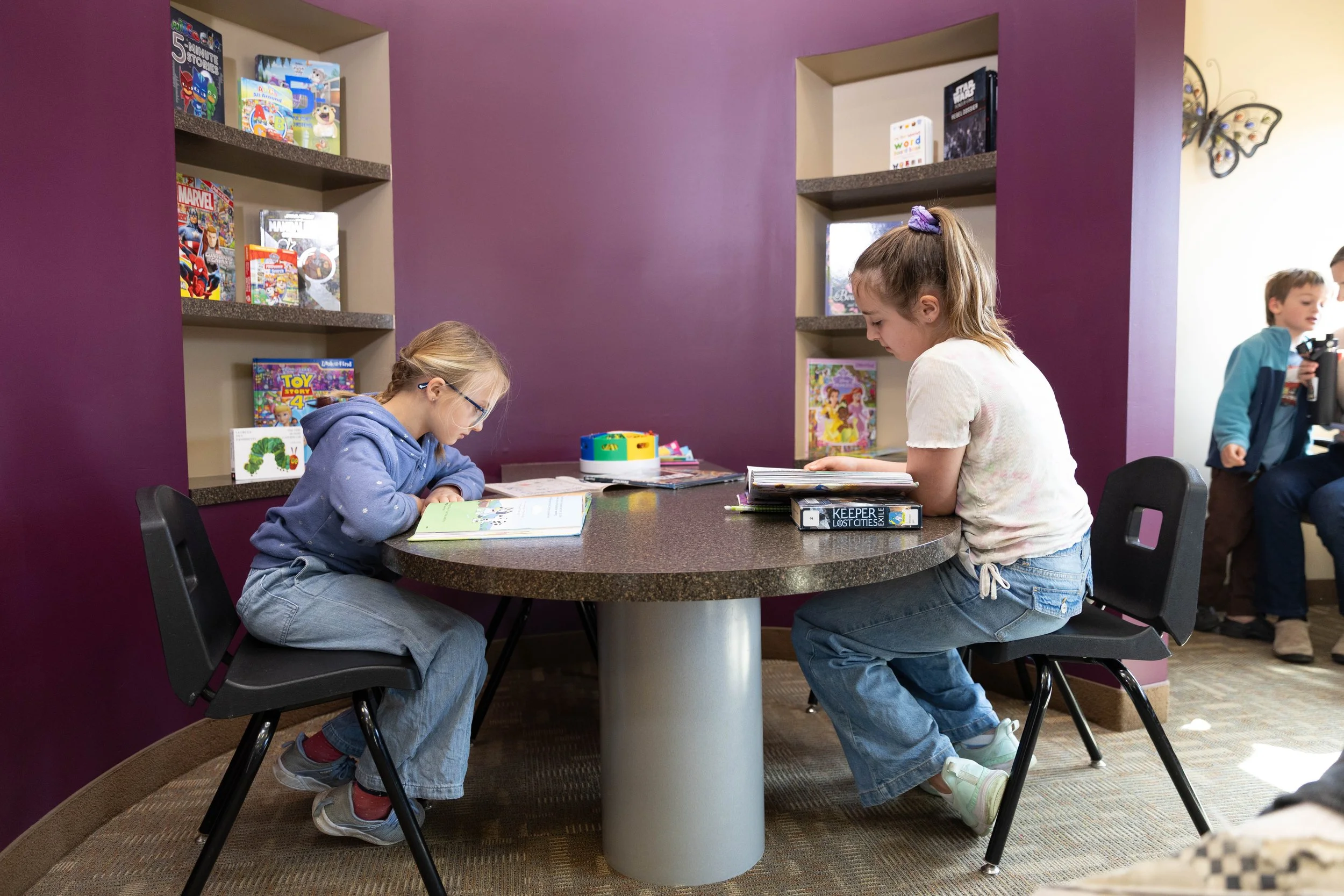 Two young girls sitting at a round table reading books in a library, with a purple wall and bookshelves behind them, and a boy and another person in the background.
