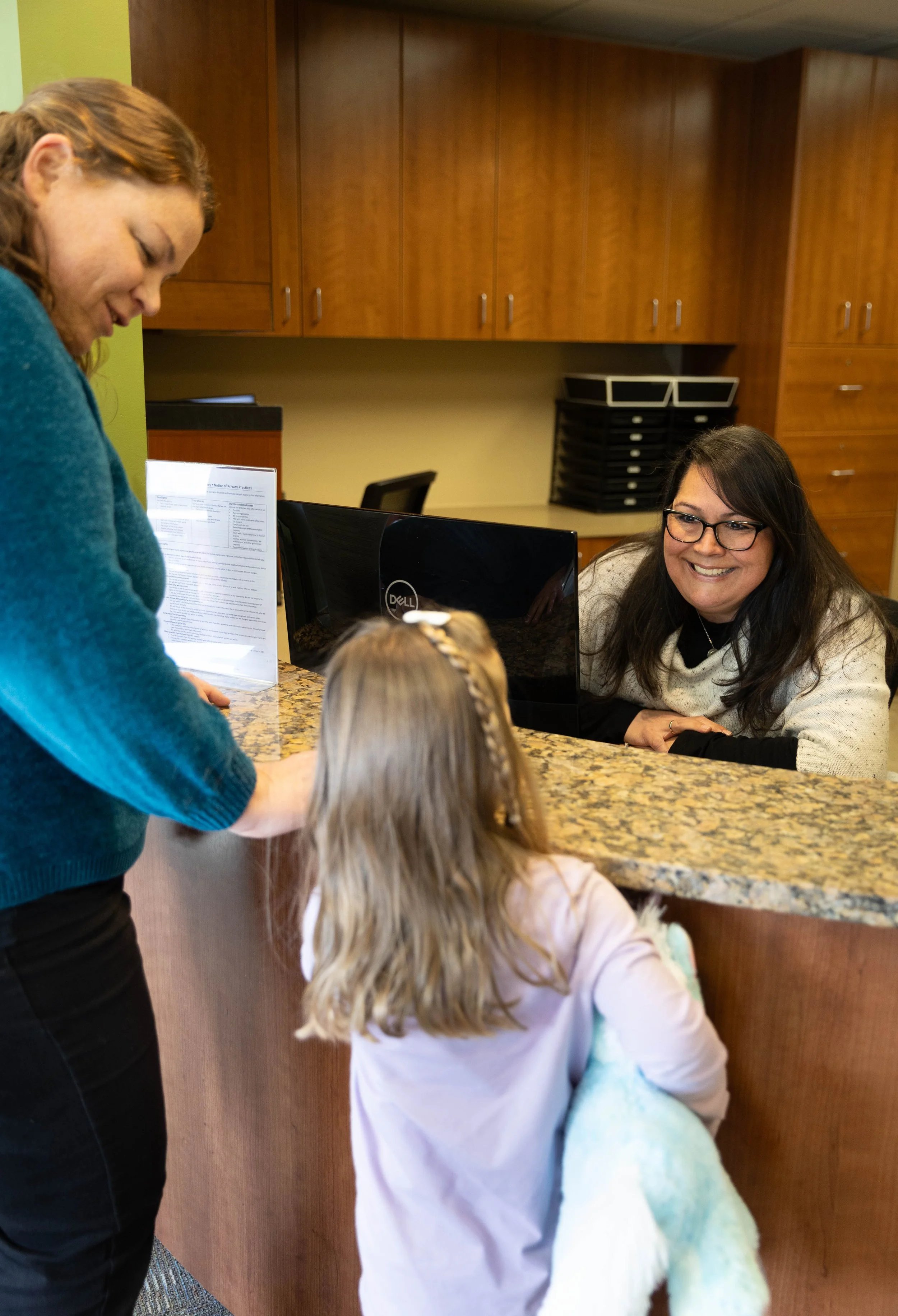A woman with glasses sitting at a reception desk talking to a child with blonde hair and another woman with red hair, in a room with wood cabinets.