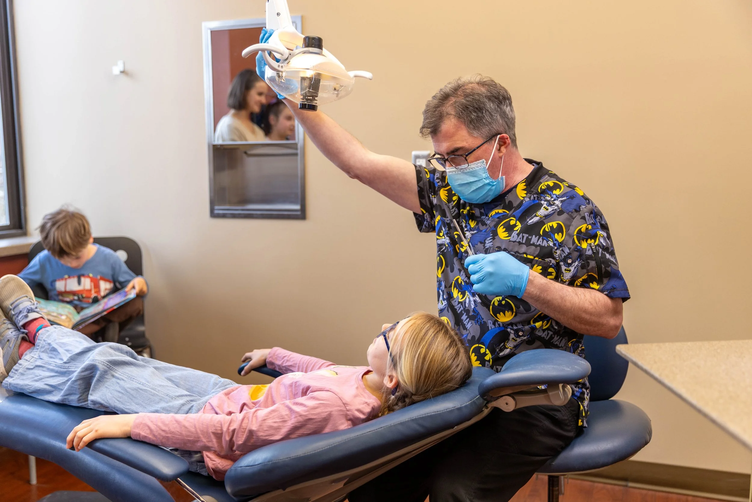 Dentist or oral health professional examining a young girl lying in a dental chair in a clinic, wearing a Batman-themed scrubs shirt, while a boy in a blue shirt reads a book in the background.