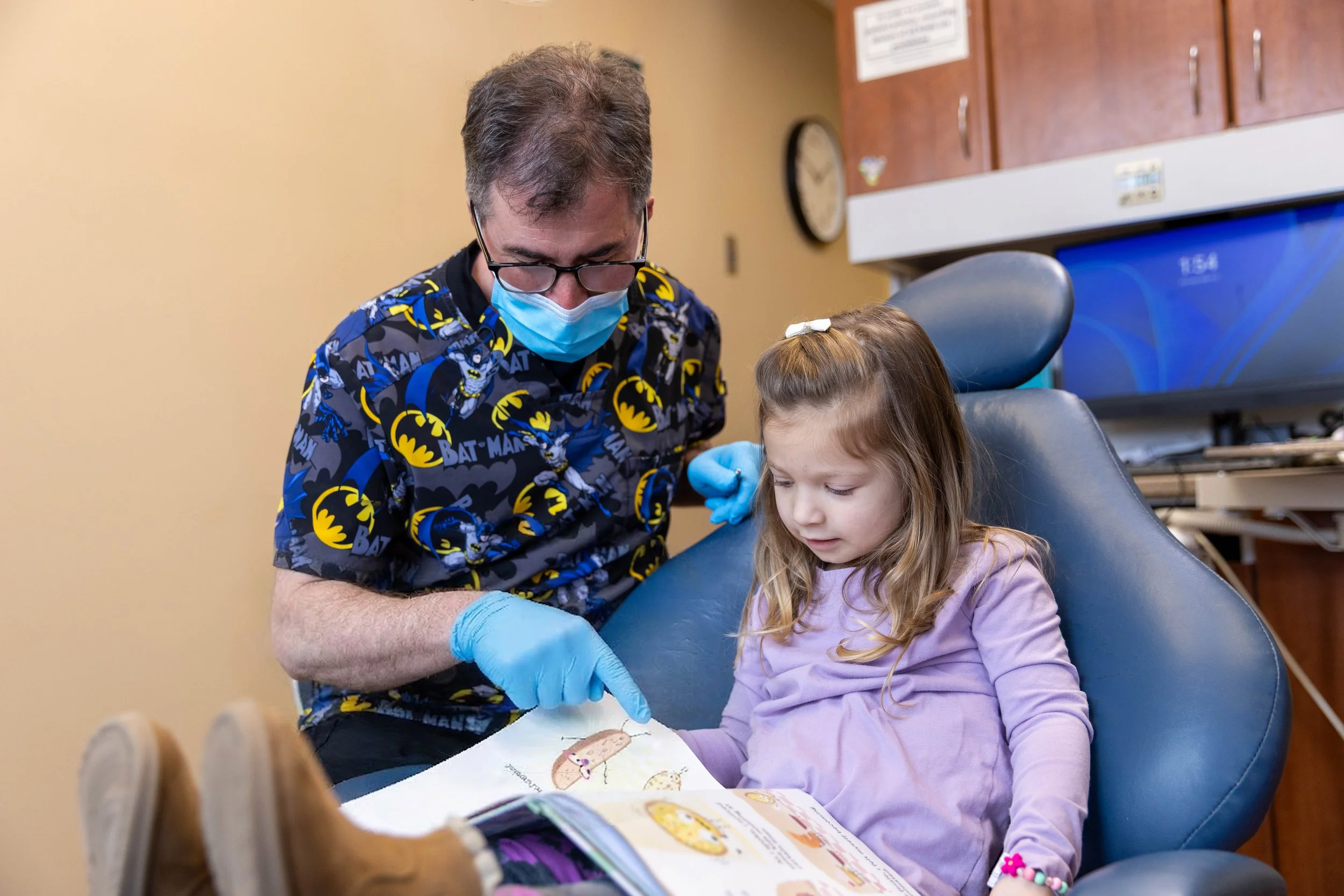 A man wearing a Batman shirt, face mask, and gloves showing a children's book to a young girl sitting in a dental chair in a dental office.