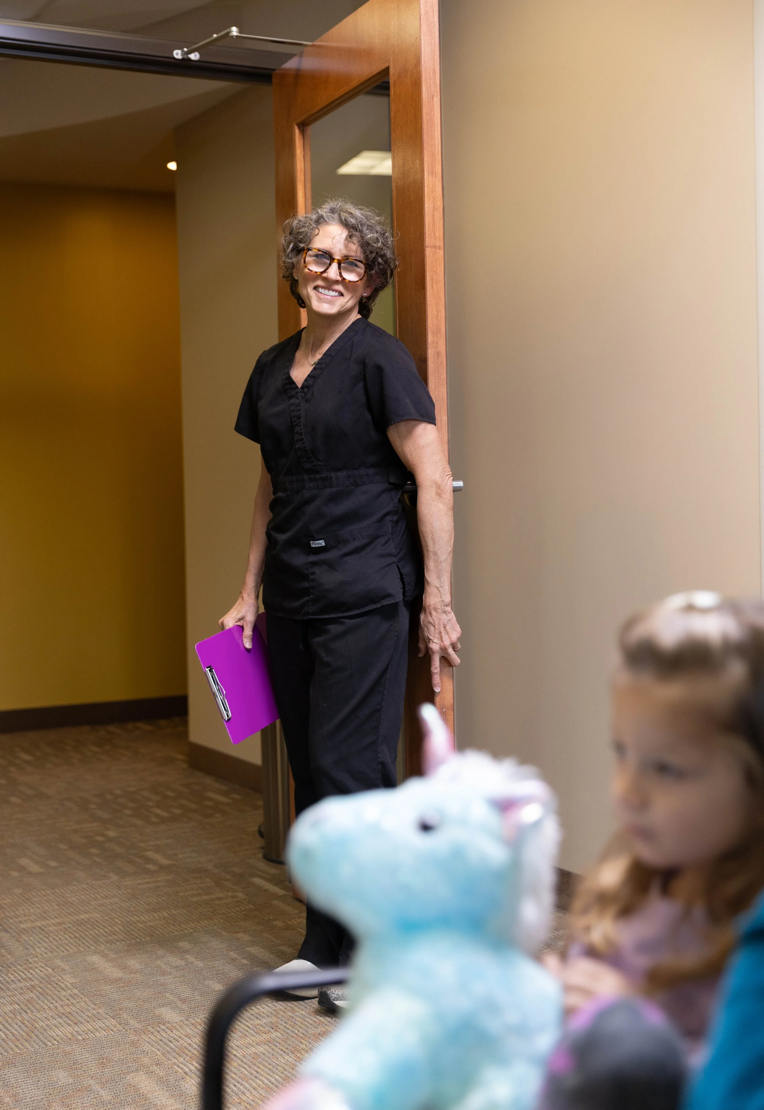 A smiling female medical professional with curly hair and glasses, wearing black scrubs, holding a purple clipboard, standing in a doorway in a medical or veterinary office. In the foreground, a young girl is playing with a stuffed unicorn toy.