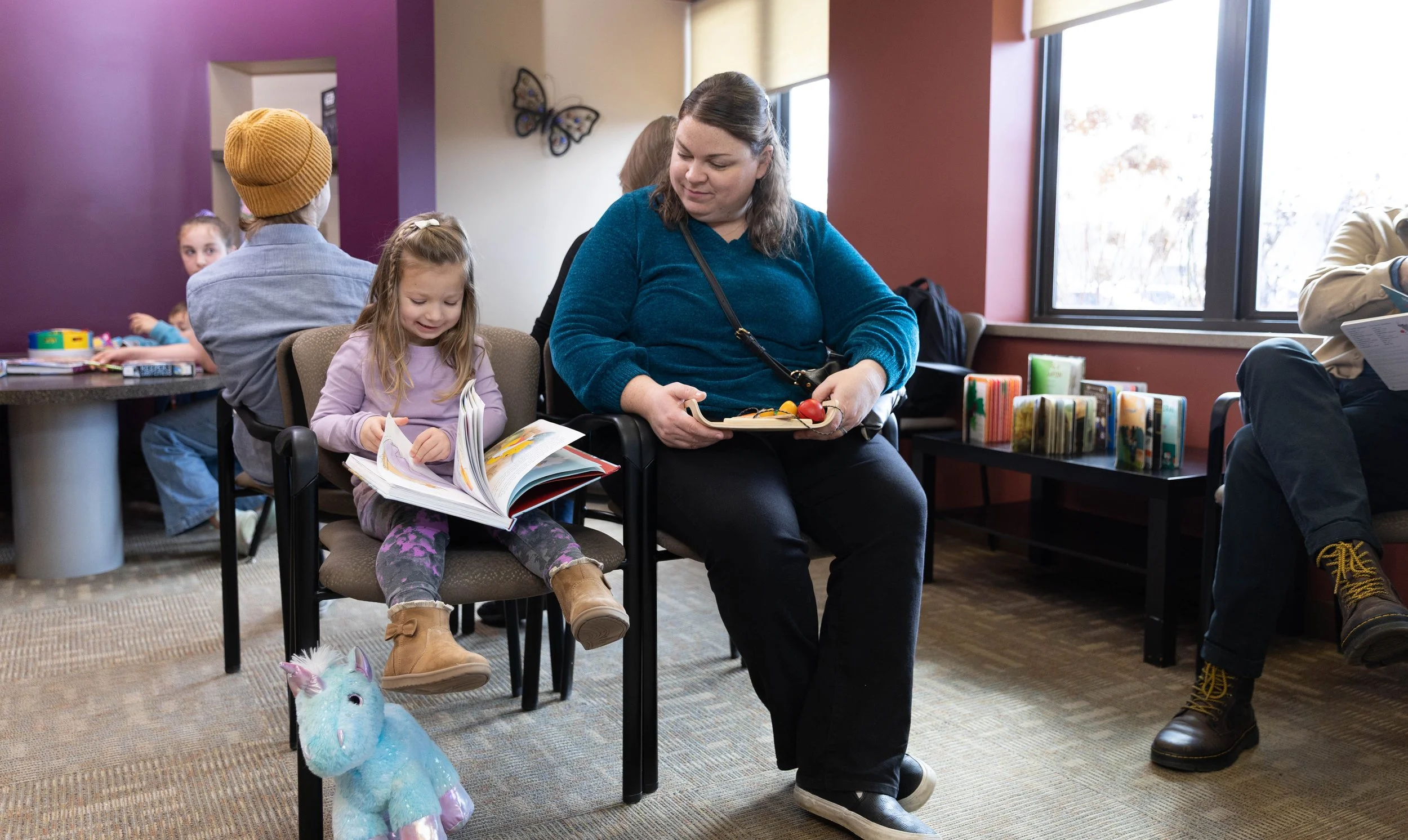 A group of people, including children and adults, sitting and reading in a waiting room with purple and beige walls, butterfly decorations, and a stack of books in the background.