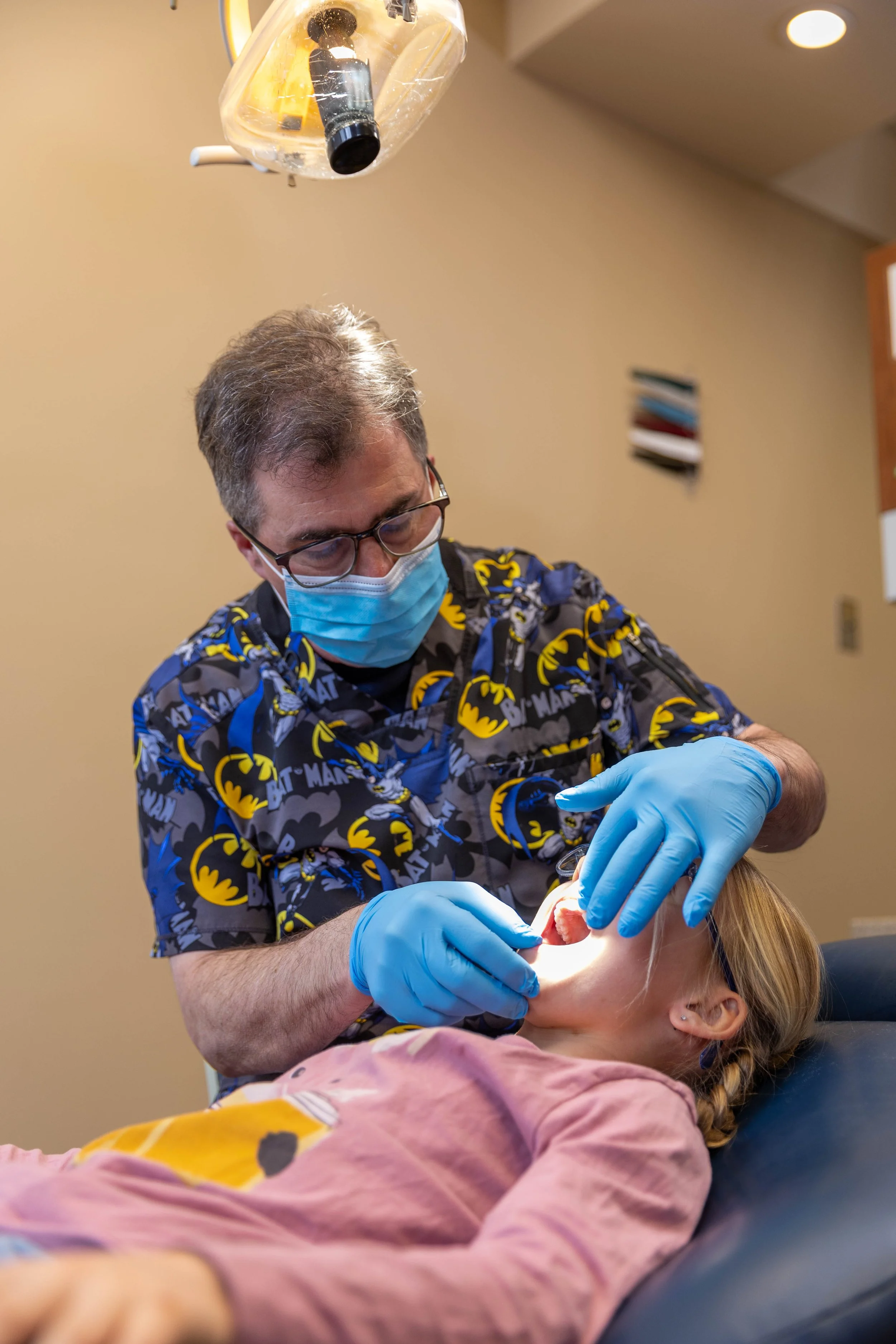 A dentist in a Batman shirt, face mask, and gloves examining a young girl lying on a dental chair in a dental office.