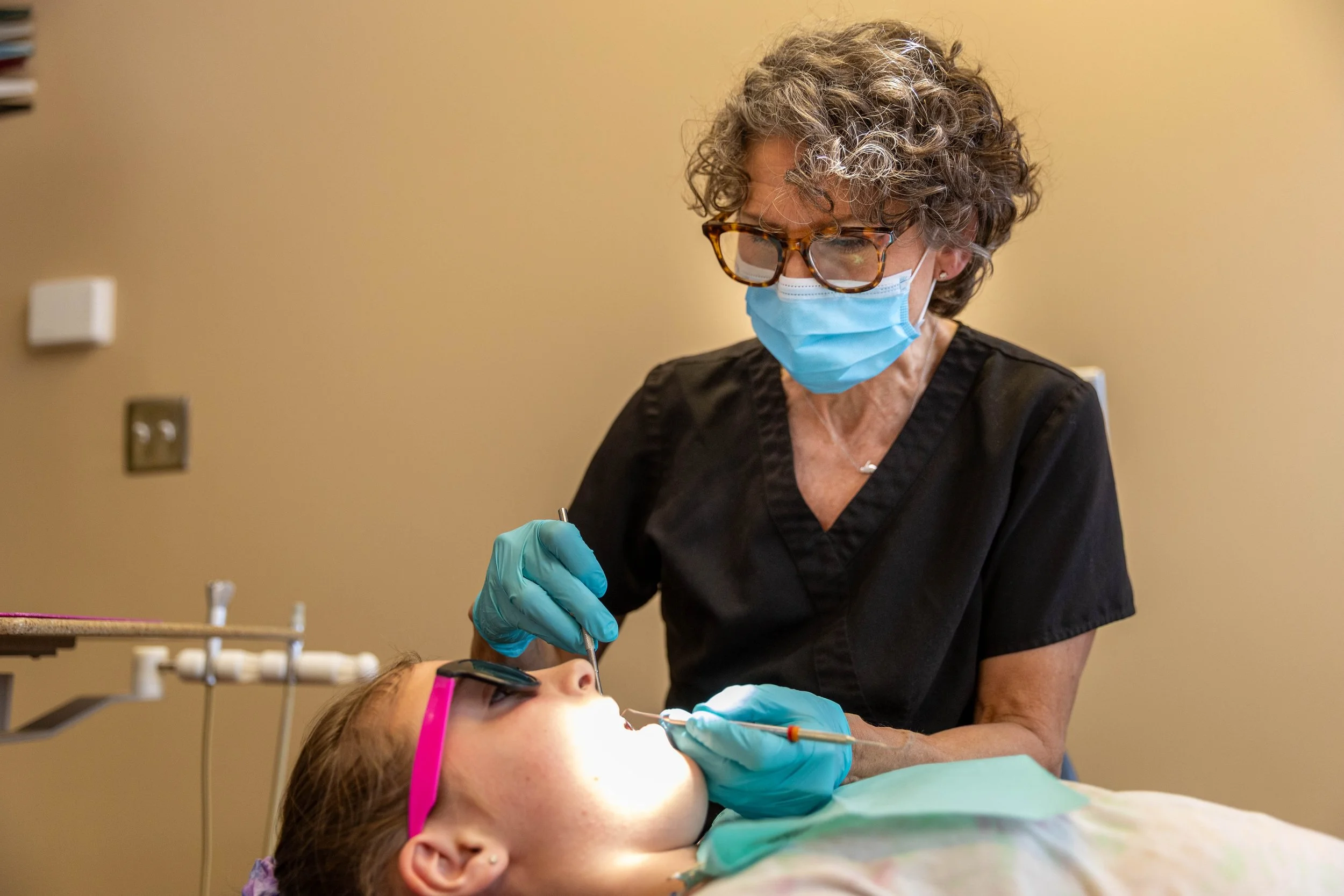 A dentist wearing glasses, a blue face mask, and teal gloves performing a dental procedure on a young patient in a dental chair. The patient is lying back, wearing pink glasses, with her mouth open.