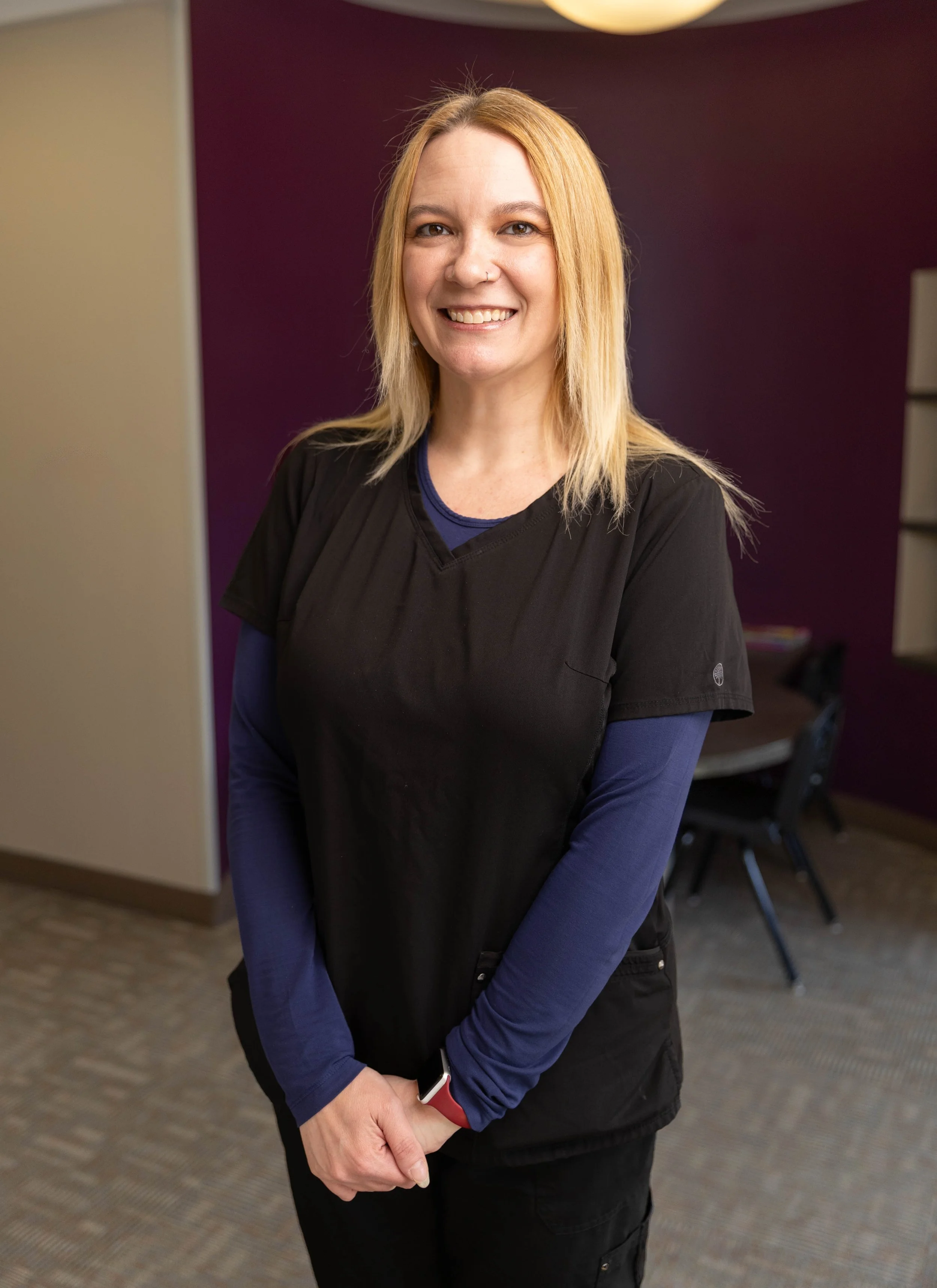 A smiling woman with blonde hair wearing medical scrubs standing indoors with a purple wall in the background.