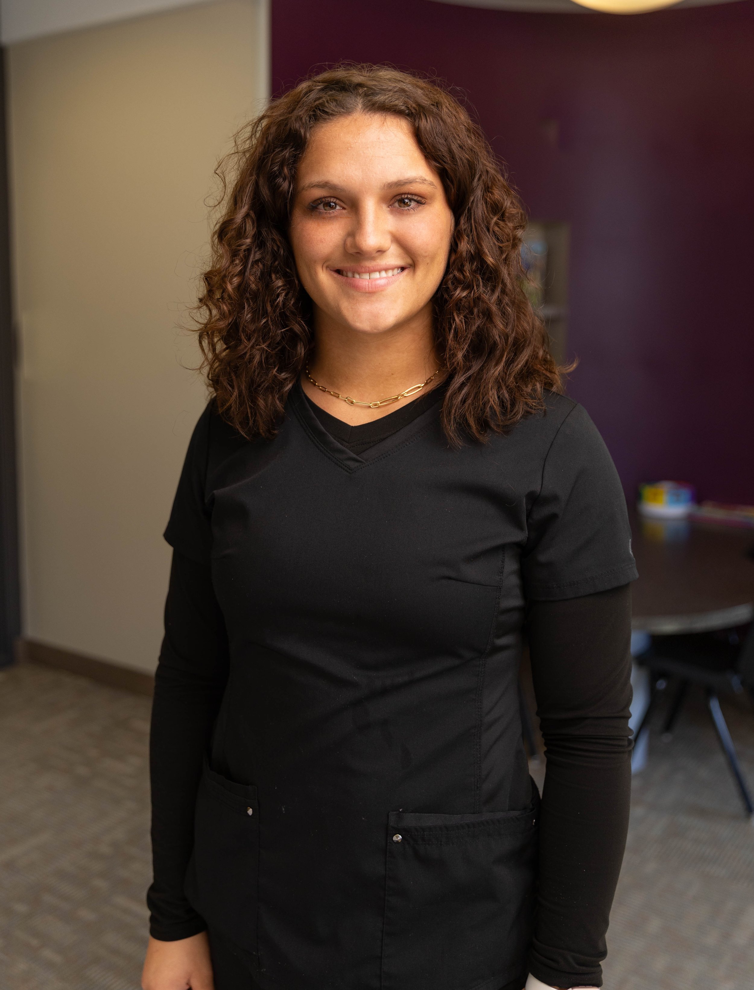 Young woman with curly brown hair smiling, wearing a black work uniform and a gold chain necklace, indoors with a dark purple wall and table in the background.