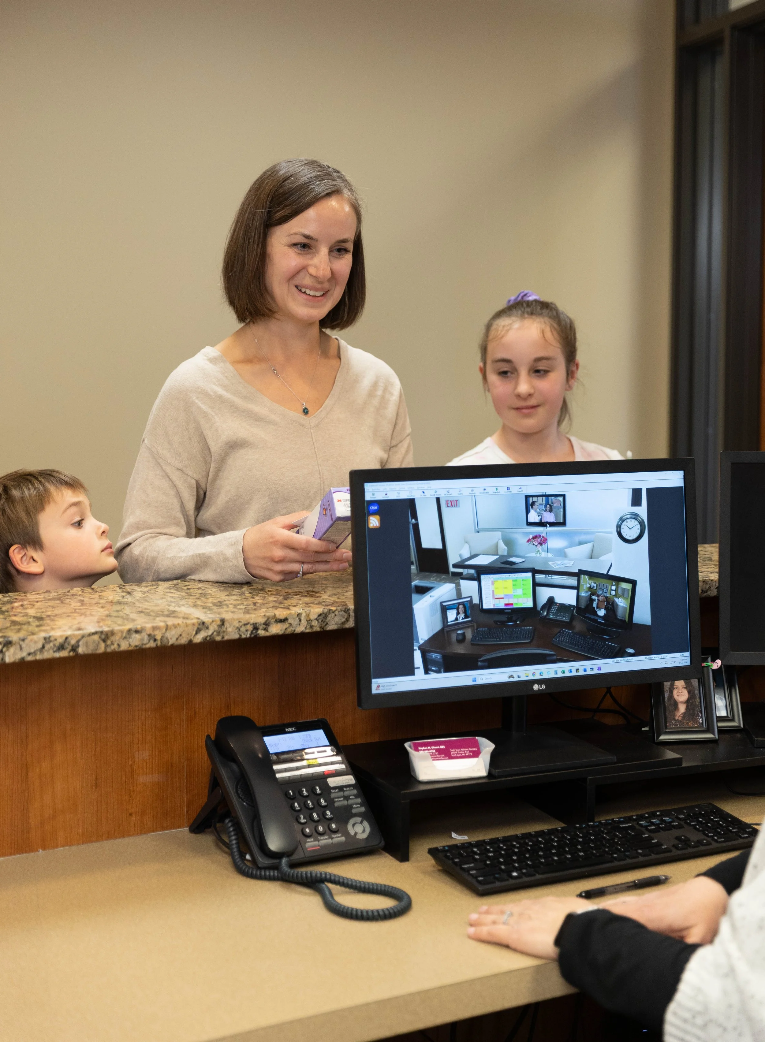A woman with two children at a reception desk, with a computer monitor displaying an office scene in the background.