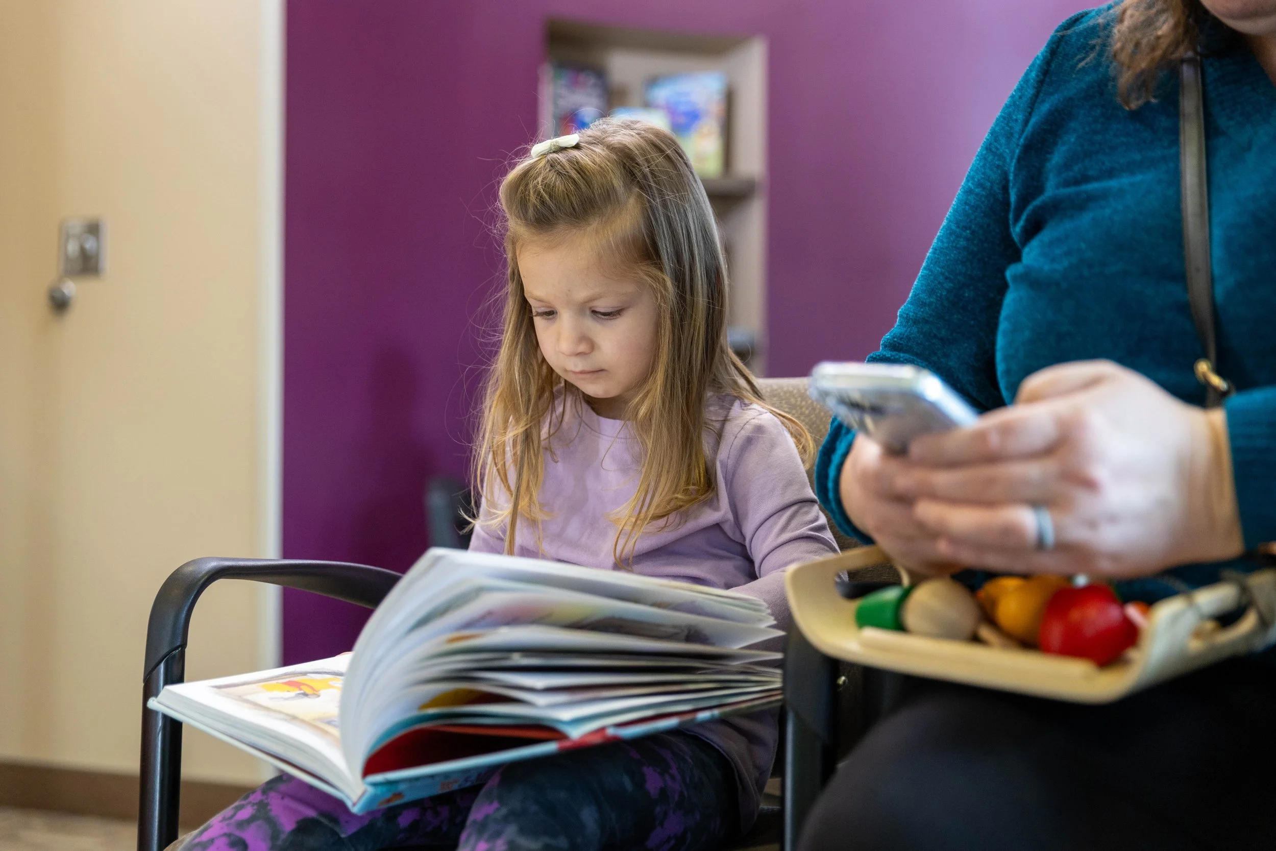Young girl with long light brown hair, wearing a purple shirt and patterned pants, sitting and looking at an open book in her lap at a waiting area.