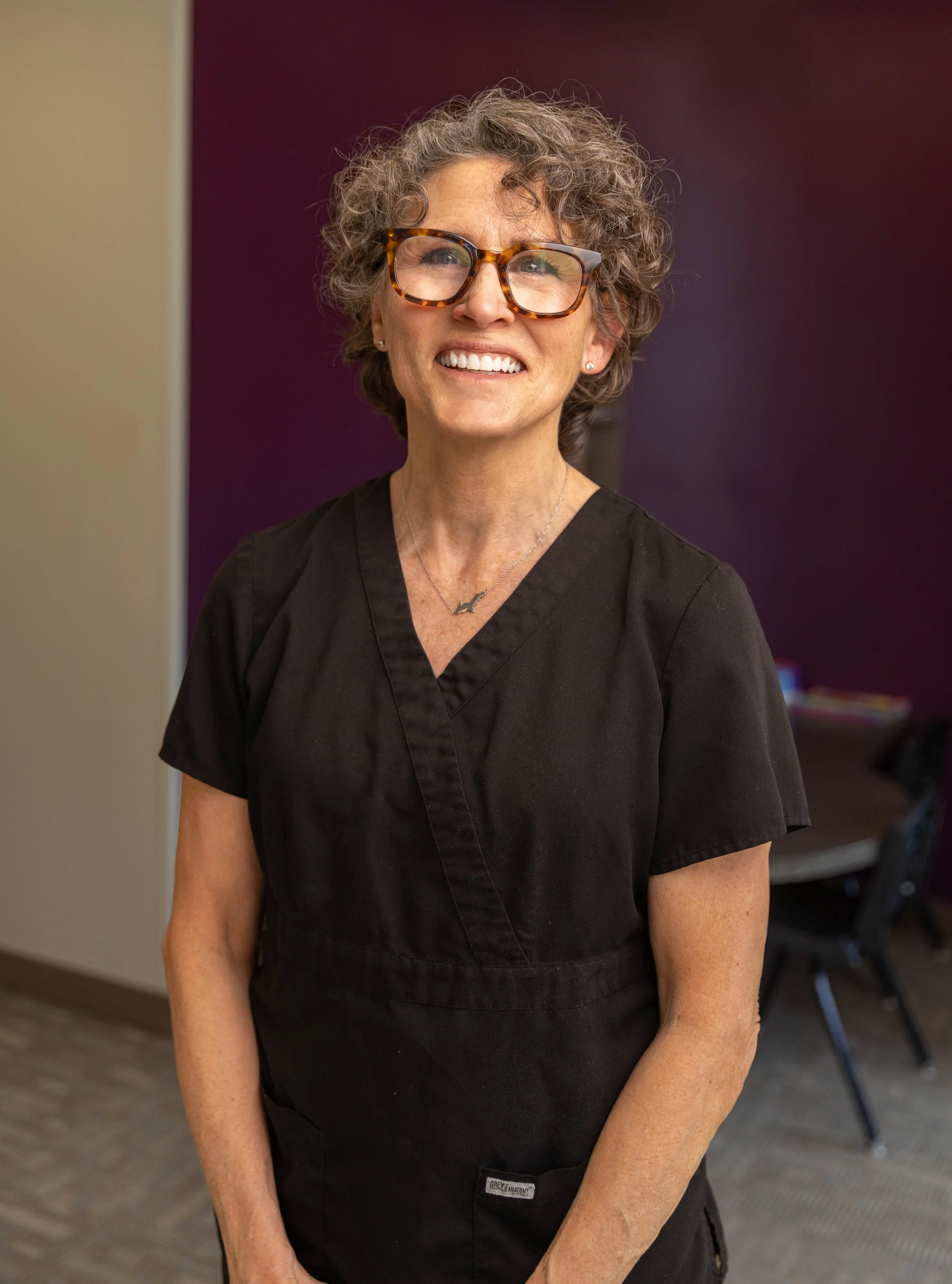 A smiling woman with curly gray hair and glasses, wearing a black medical scrub top, standing in an indoor setting with a purple wall and chairs in the background.