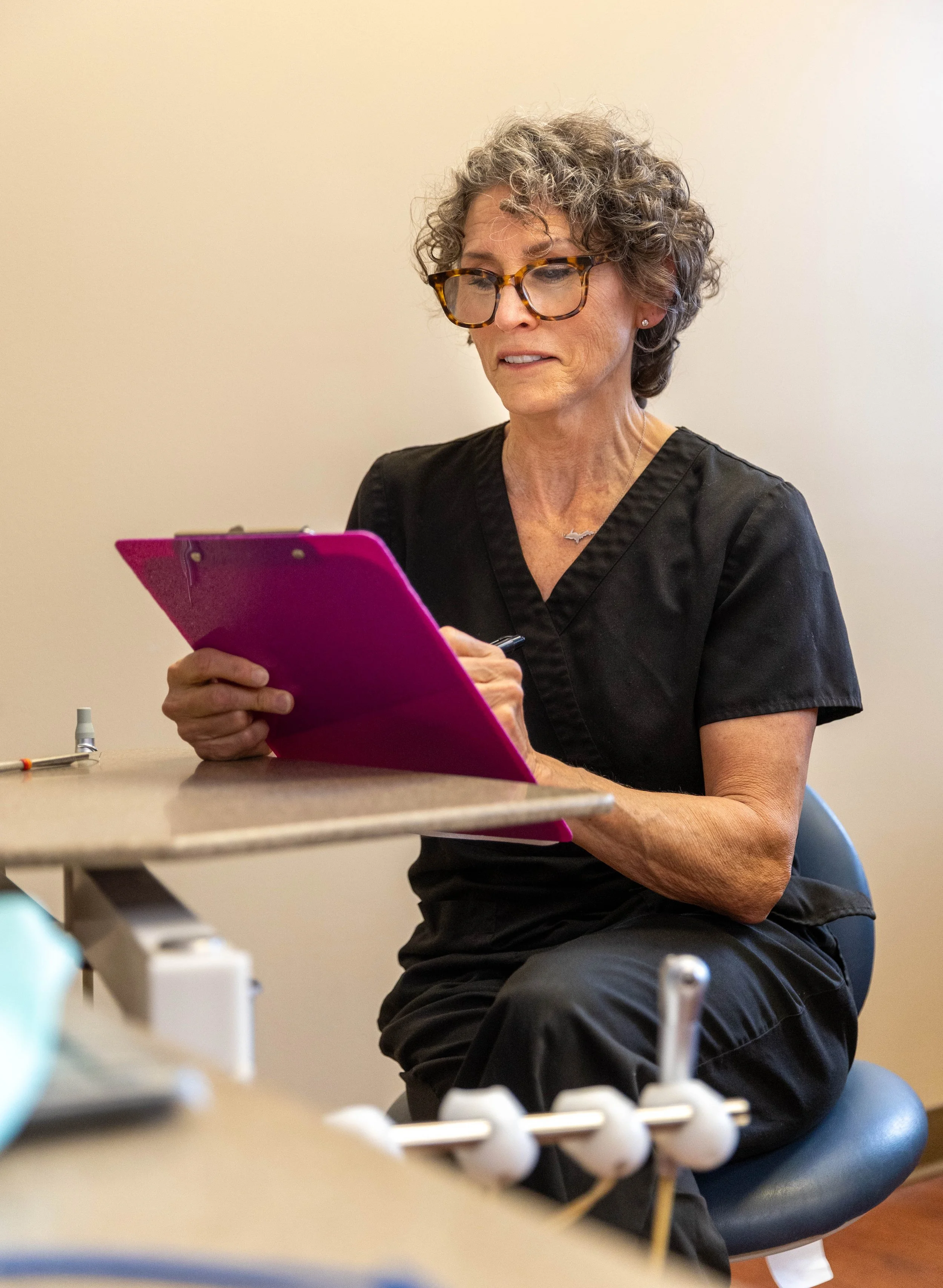 A woman with curly gray hair, glasses, and wearing black scrubs, sitting on a stool and looking at a pink clipboard in her hands.