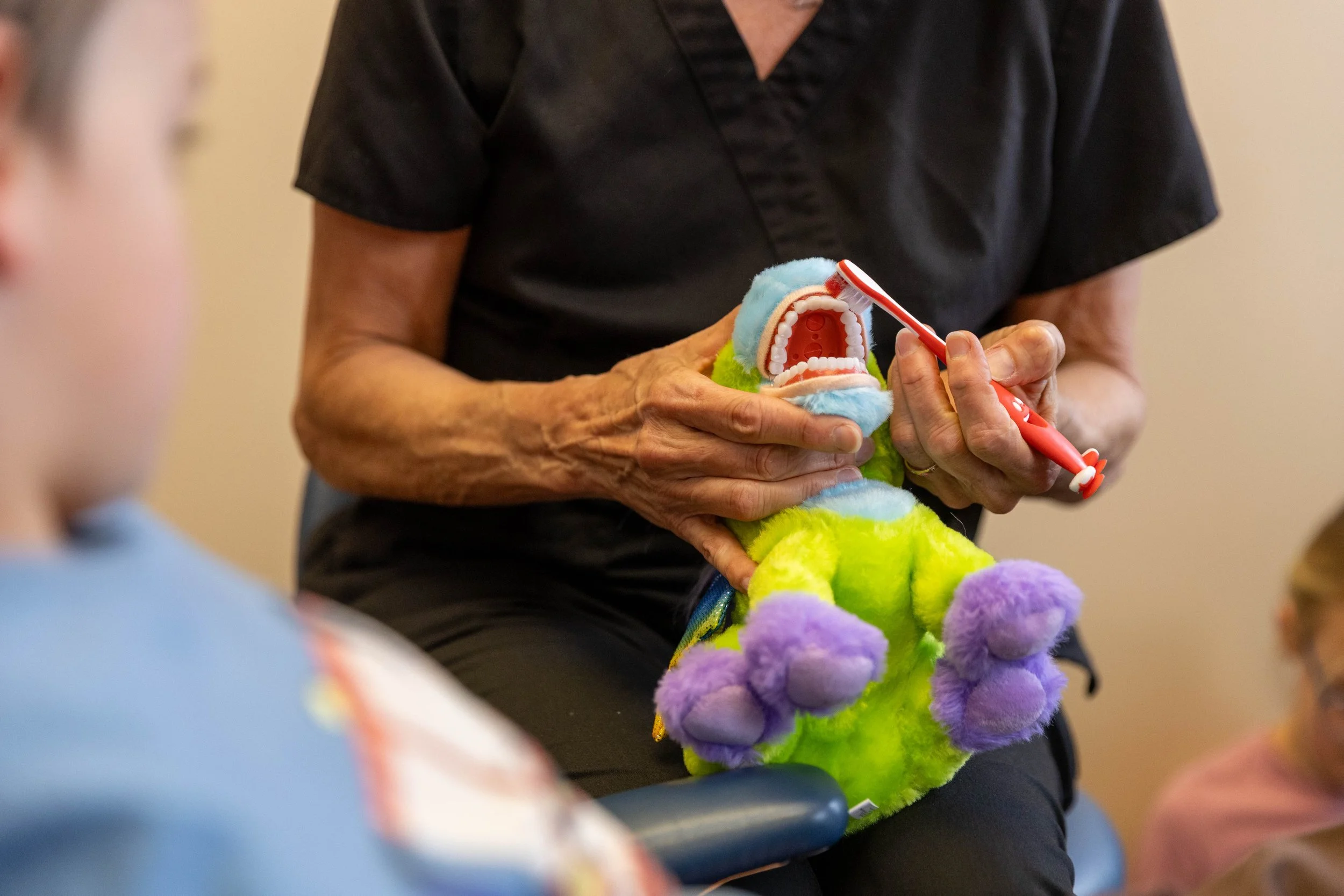 A person demonstrates brushing the teeth of a colorful stuffed plush toy with a red toothbrush. The person is wearing a black uniform, and the plush toy has vibrant blue, green, purple, and yellow colors.