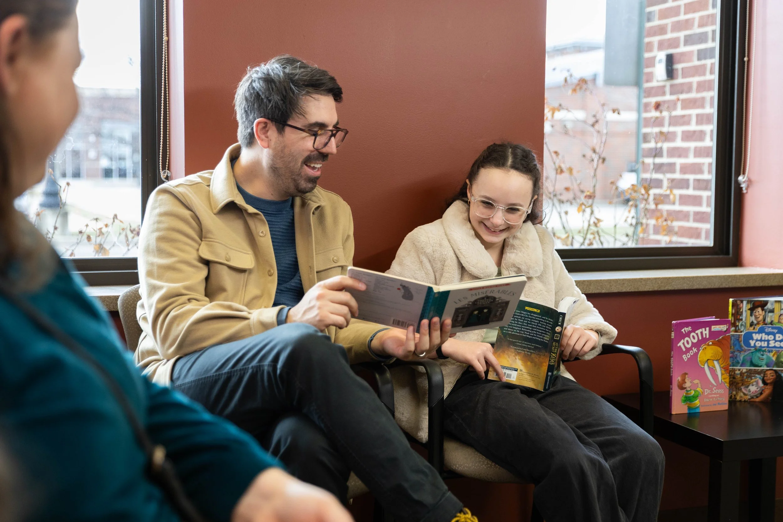 A man and a girl sitting on chairs reading books while another person sits nearby. The girl is smiling and wearing glasses, holding a book, while the man, wearing glasses, is also engaged in reading. There are more books on a table next to them.