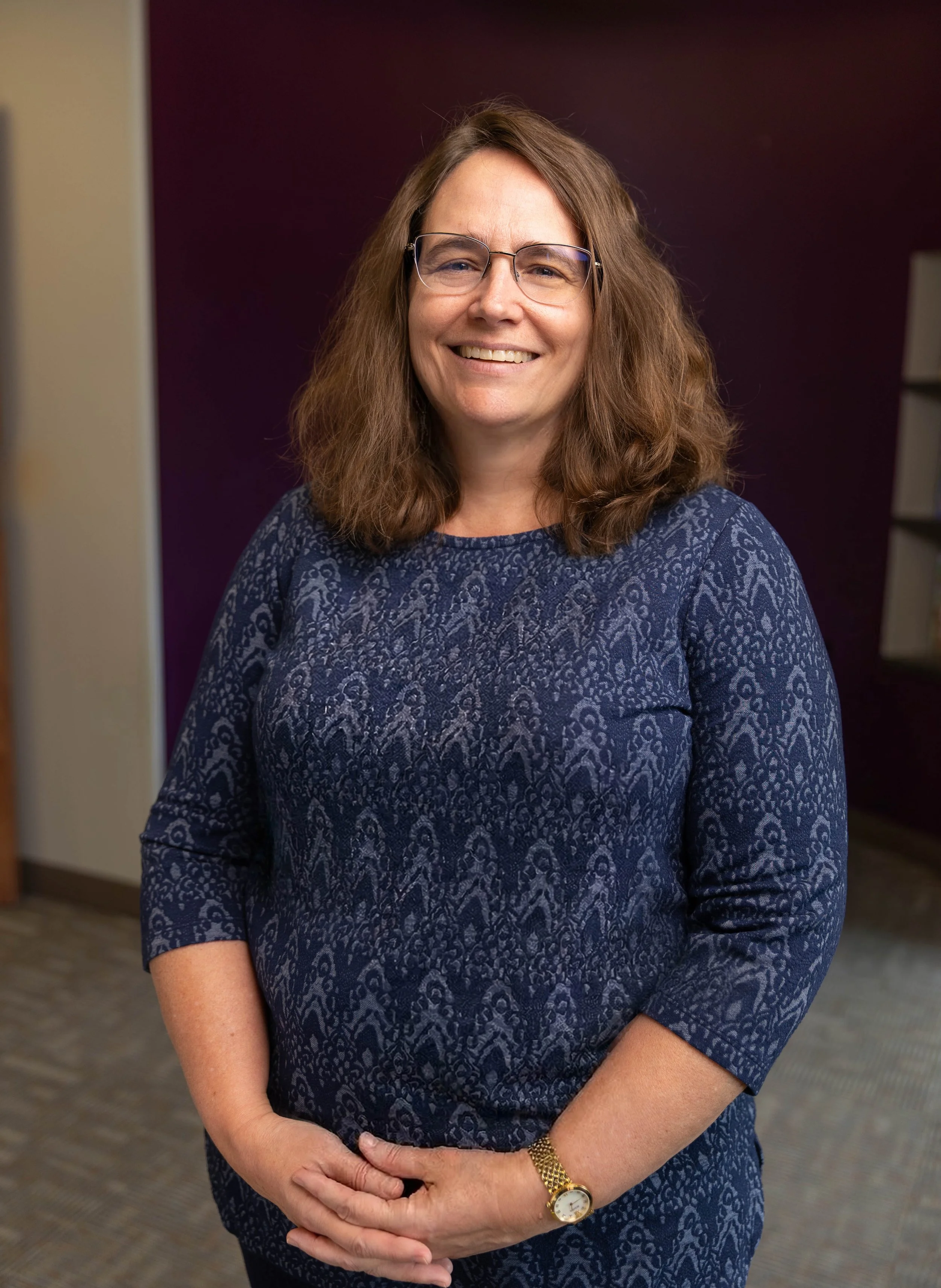 A smiling woman with shoulder-length brown hair, wearing glasses, a blue patterned dress, and a gold watch, standing indoors against a dark background.