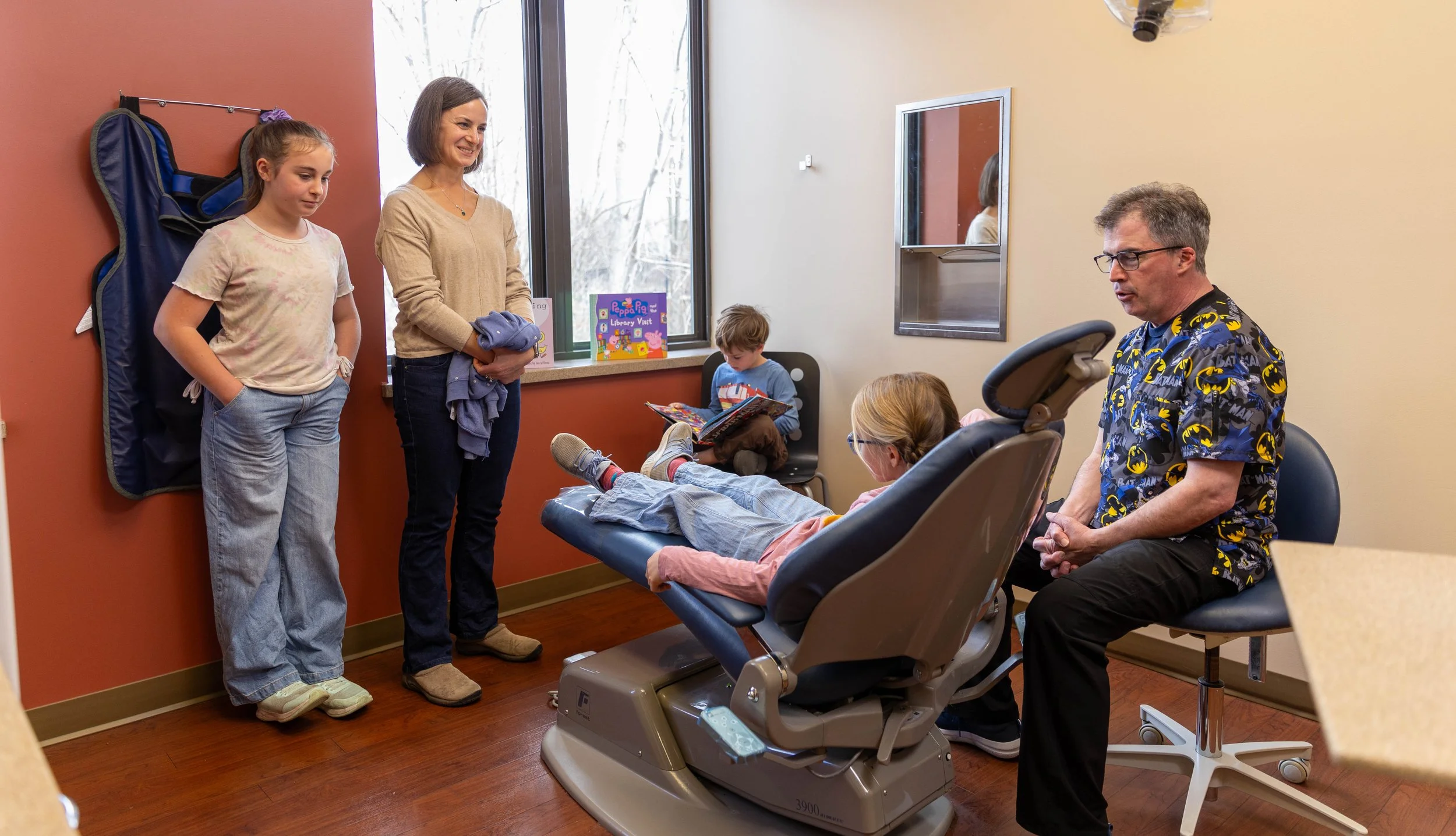 Child lying in a hospital bed with a woman sitting next to her and children standing nearby, in a medical examination room.