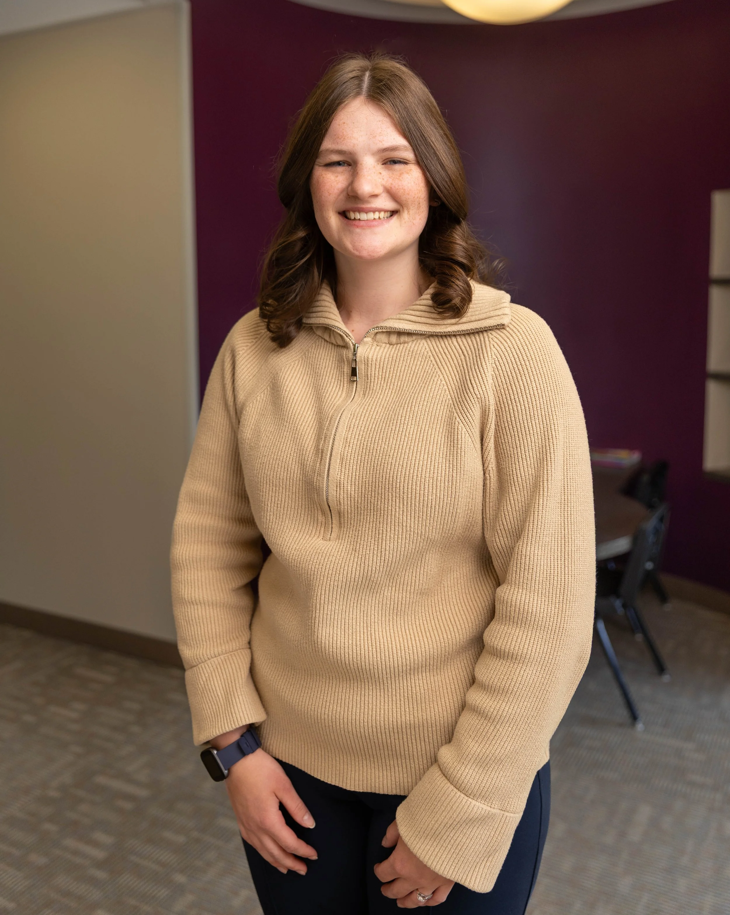 A young woman with shoulder-length brown hair, smiling, wearing a beige zip-up sweater and dark pants, standing indoors against a purple wall and beige partition.