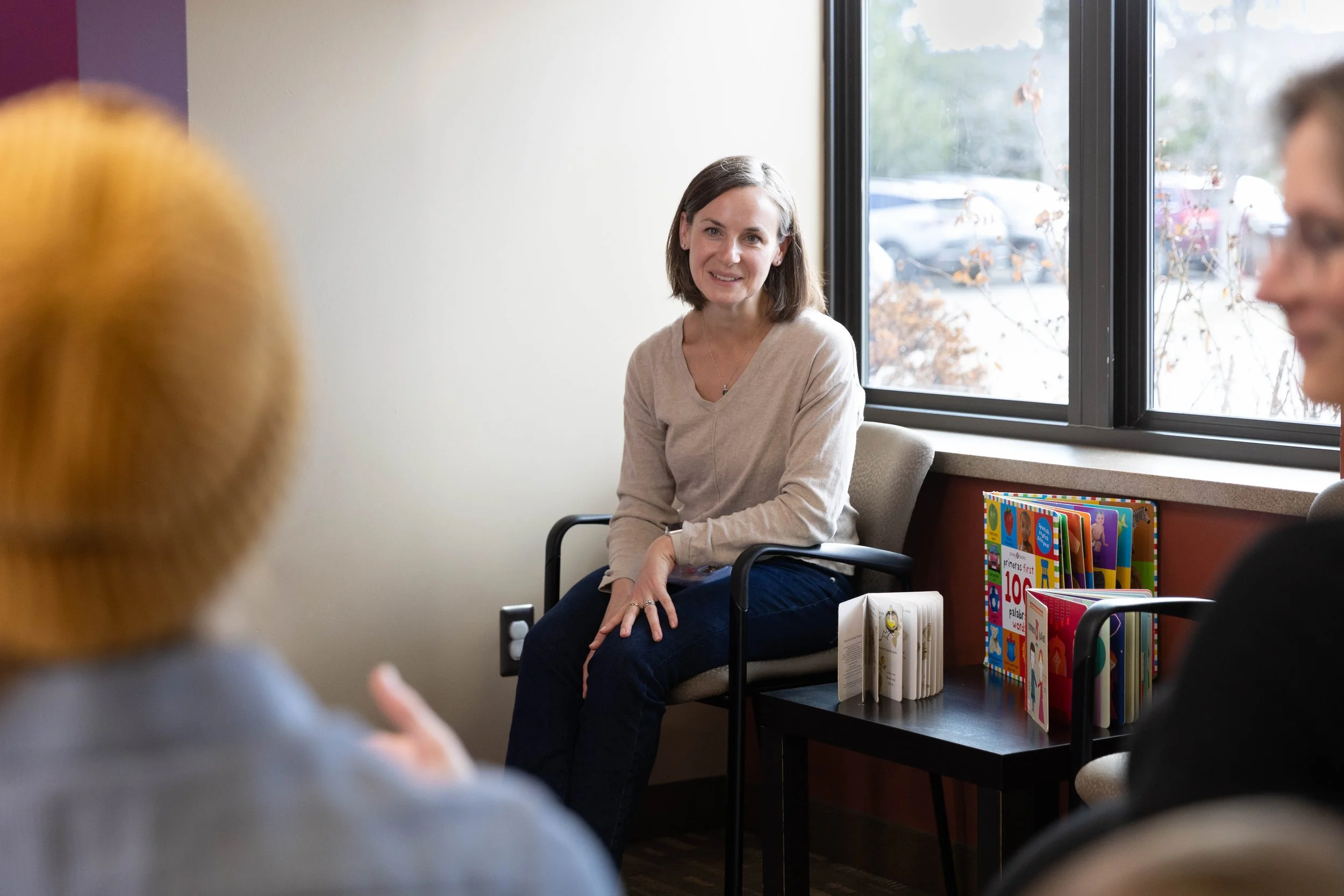 A woman with shoulder-length brown hair and a beige sweater sitting on a gray office chair, engaged in conversation with two blurred individuals in a room with a window showcasing a parking lot outside. There are children's books on a black table bes