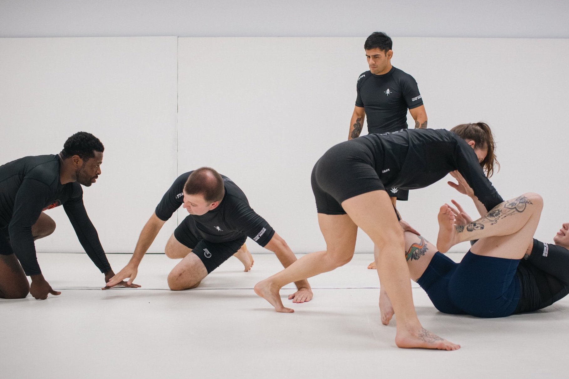 Mixed martial arts training session with women and men practicing grappling on a white mat in a gym.
