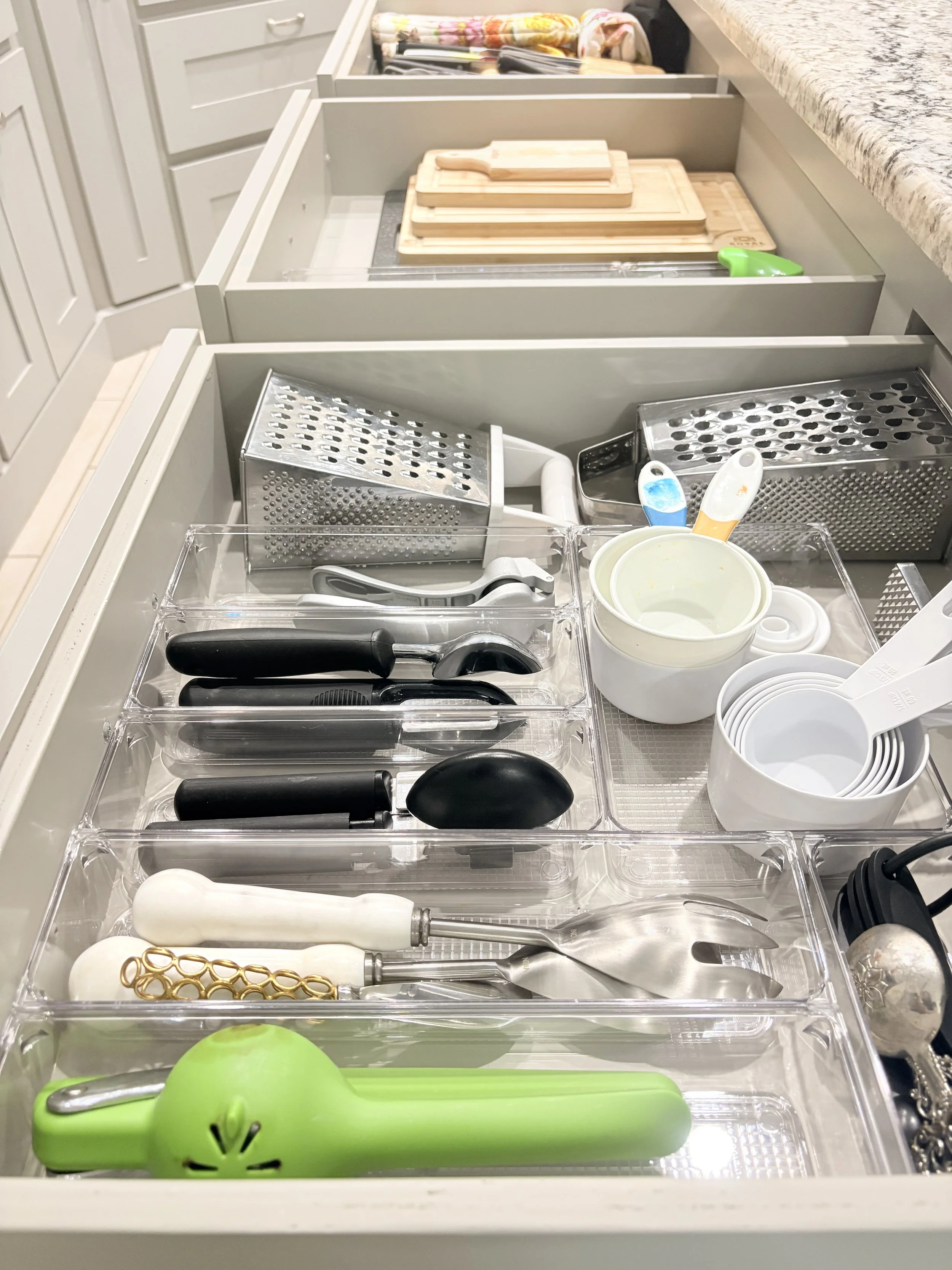Drawer filled with kitchen utensils and tools, including a green garlic peeler, measuring cups, a can opener, knives, graters, and cutting boards.