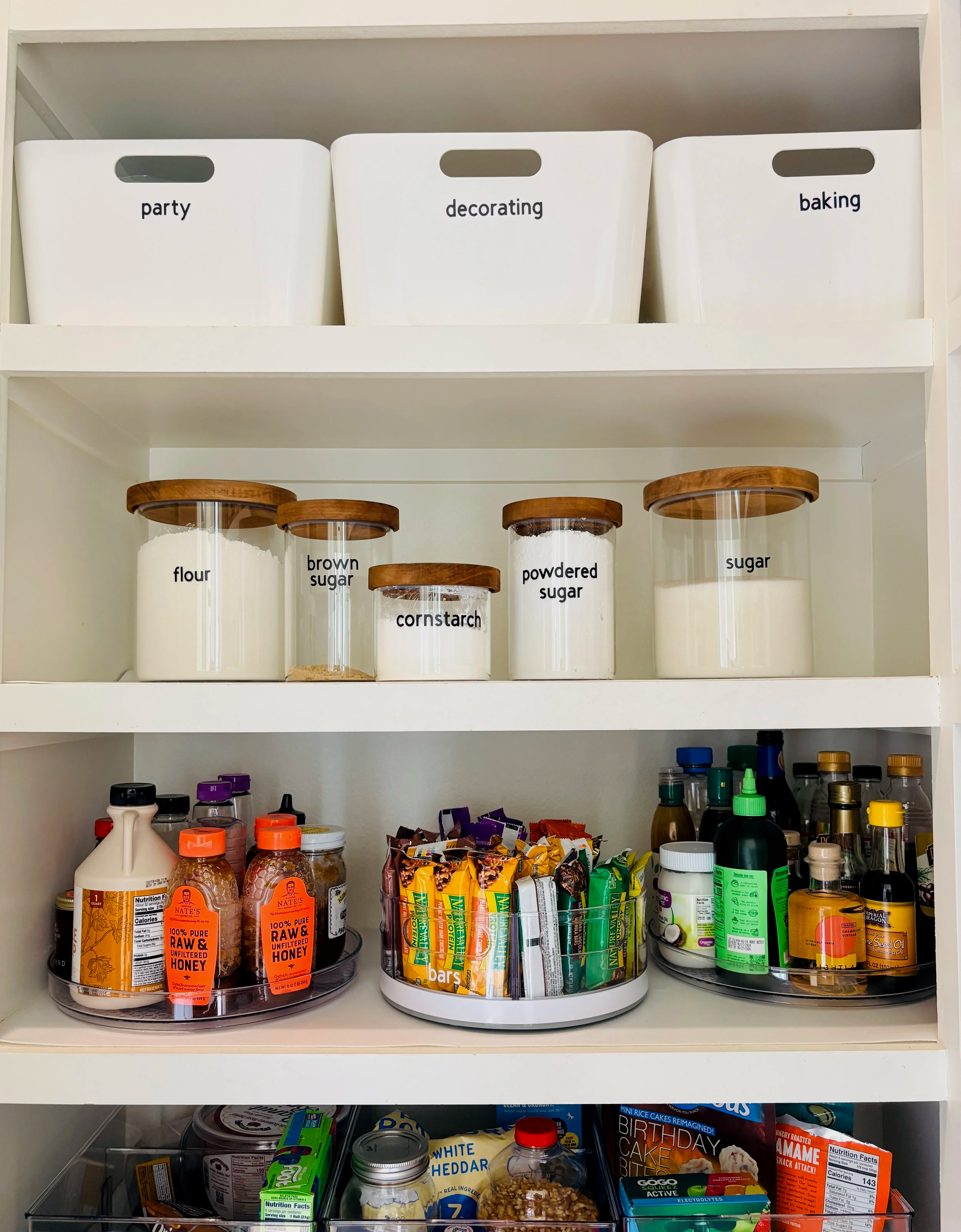 Shelf with labeled storage bins for party, decorating, and baking supplies. Lower shelves contain various food items, snacks, condiments, and baking ingredients.