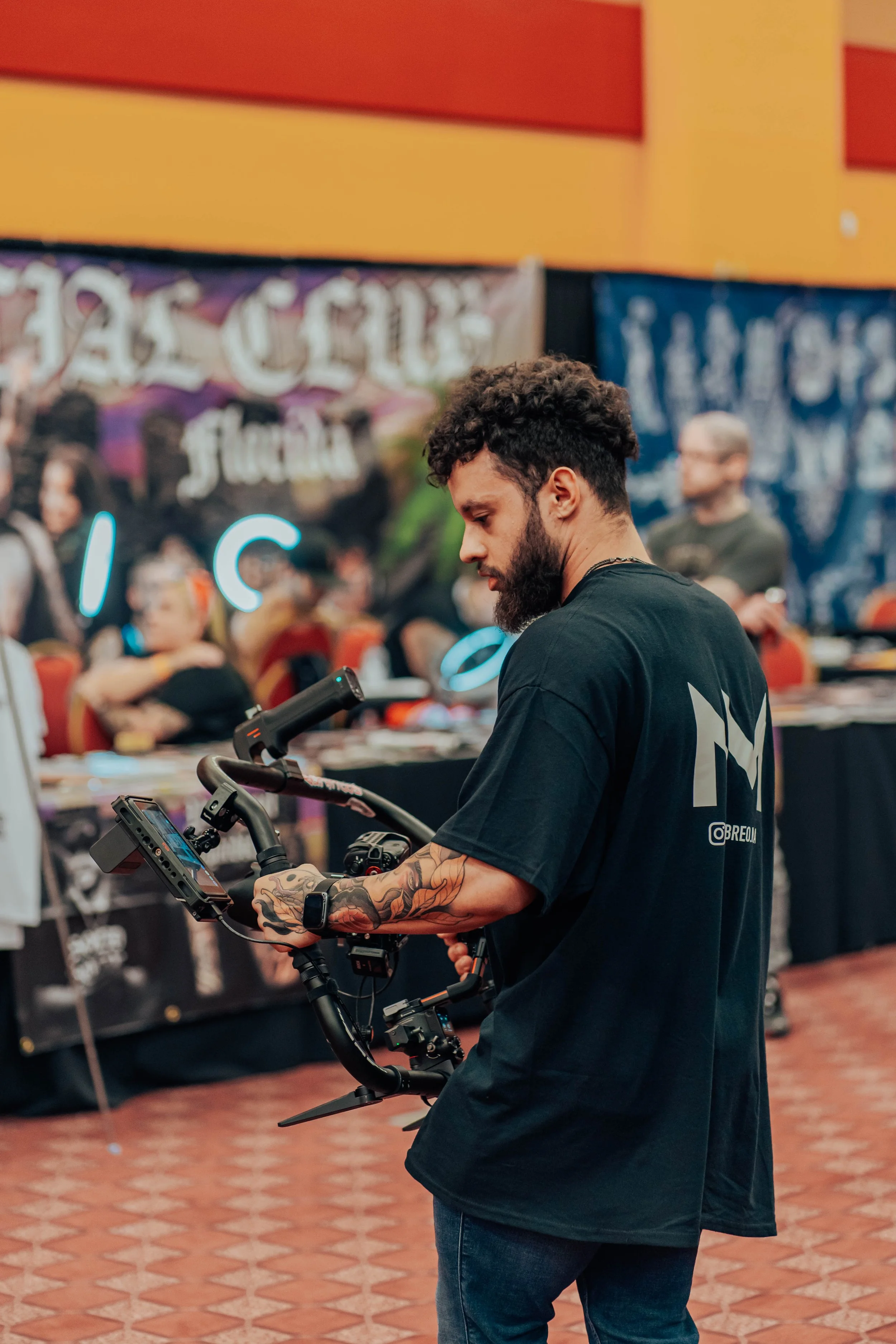 A man with a beard and tattoos on his arms operating a handheld camera gimbal in a crowded indoor event space.