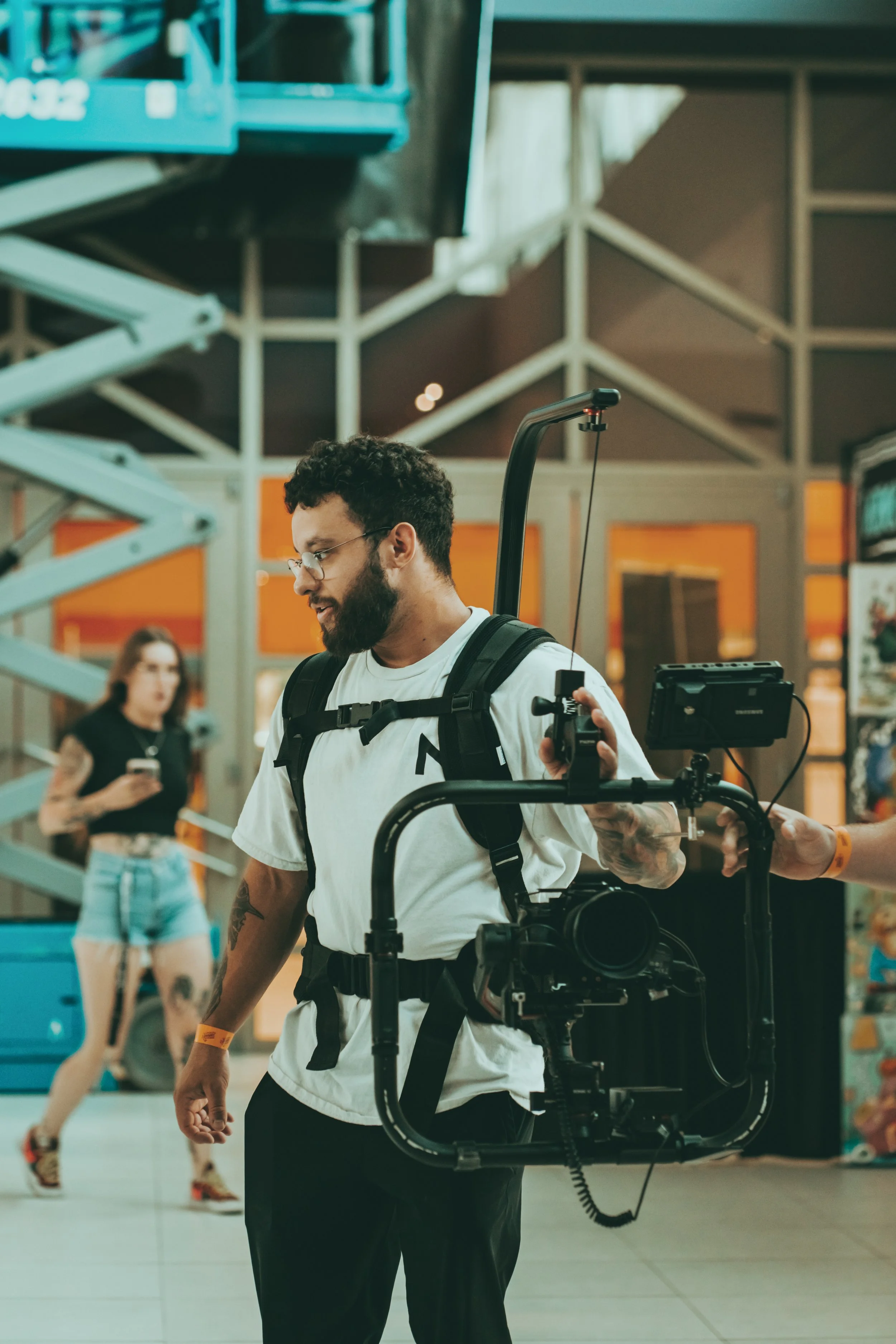 A man with glasses, a beard, and a white t-shirt carrying a camera rig, standing in an indoor public space with a woman in the background using her phone.