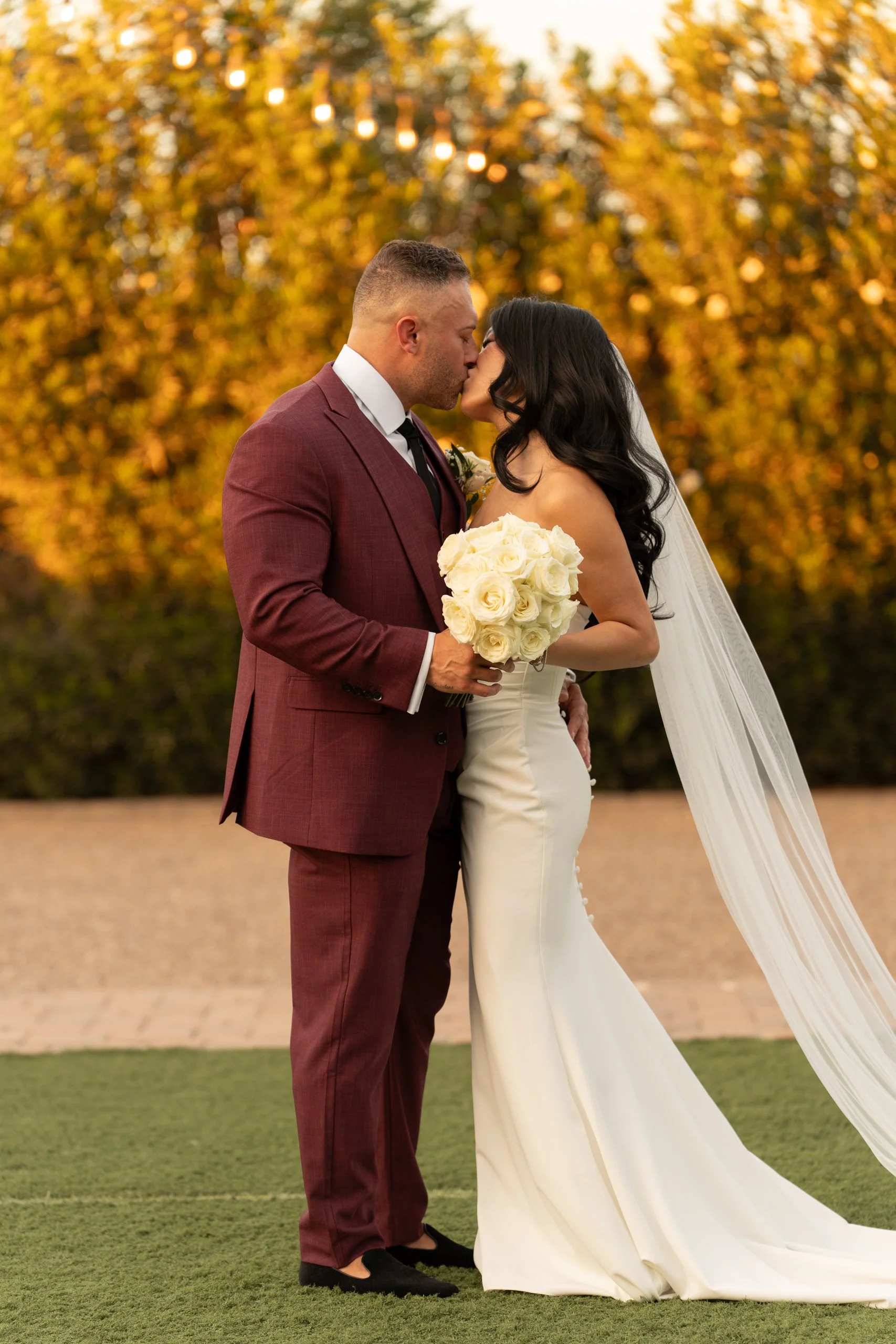 A bride and groom kiss outdoors during sunset. The bride has long black hair, wears a white strapless wedding gown with buttons down the back and a veil, and holds a bouquet of white roses. The groom is dressed in a maroon suit, white shirt, and black shoes. The background features autumn-colored trees and warm lighting.