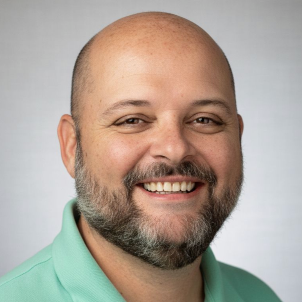 A man with glasses, a bald head, and a goatee, wearing a suit and tie, smiling in front of an abstract red and orange background.
