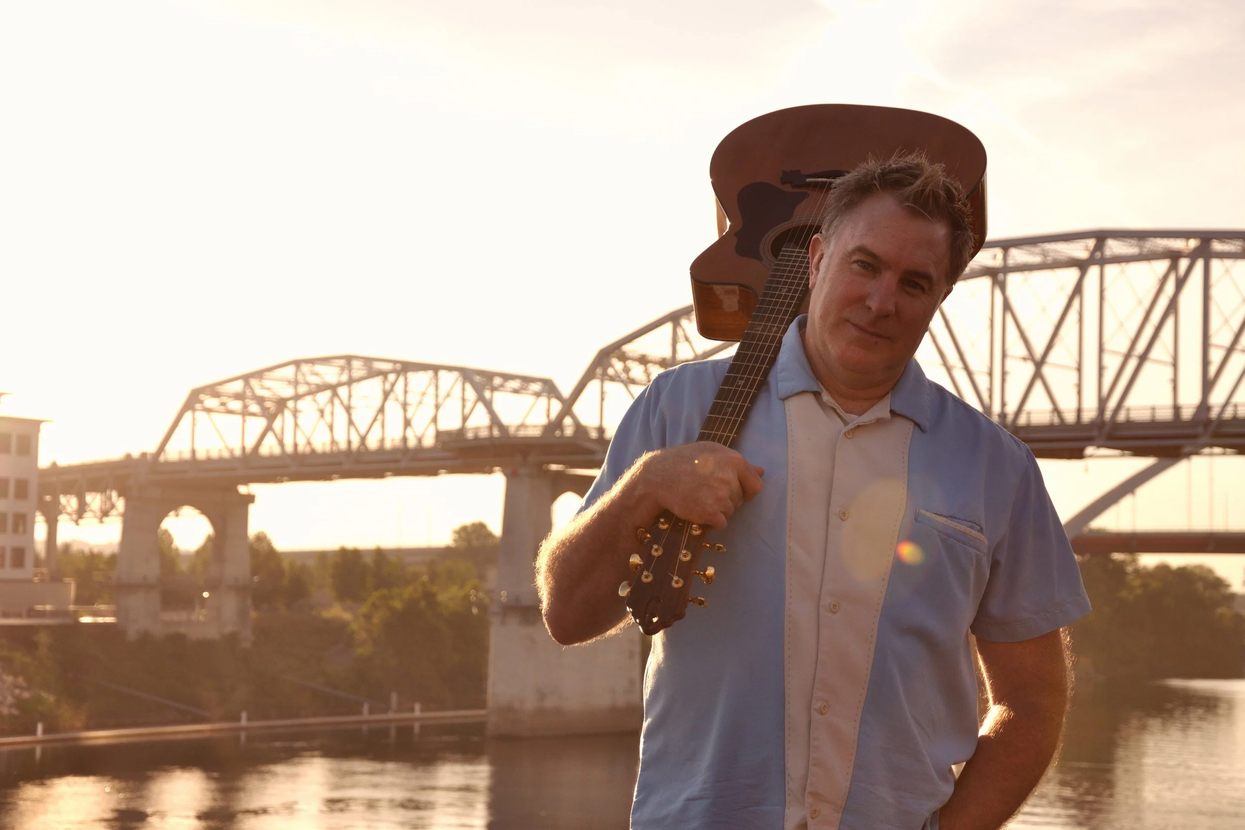 Shane Adams with a guitar over his shoulder by a Nashville river bridge at sunset