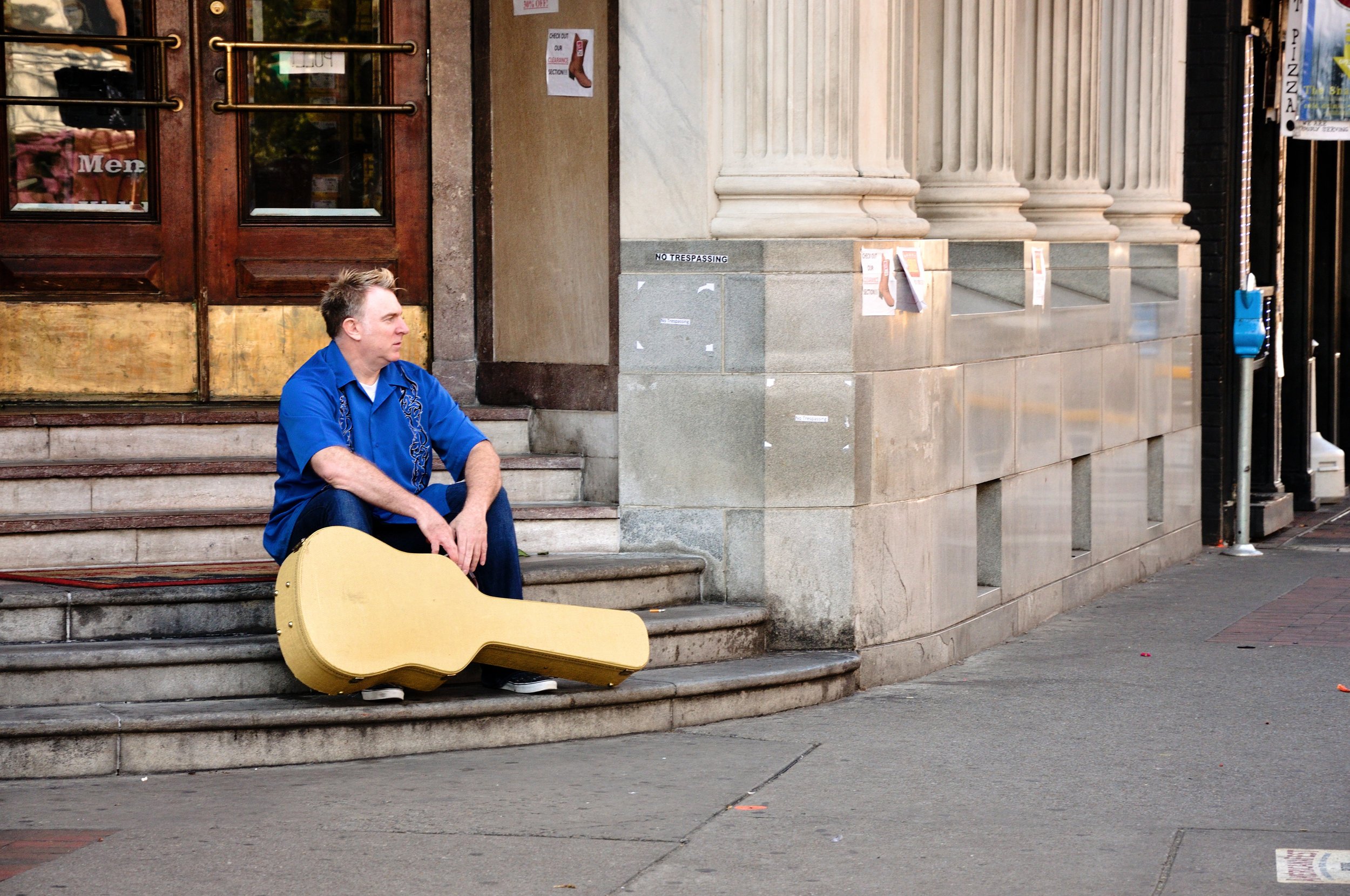 Shane Adams sitting on downtown steps with a guitar case