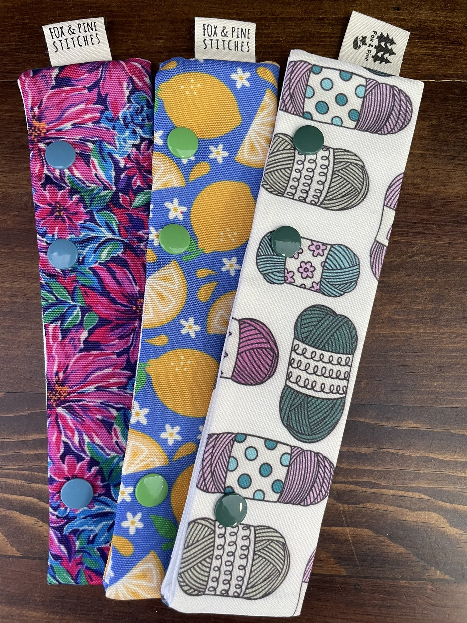 A variety of Fox & Pine double-pointed needle holders in bright designs, including floral, lemon, yarn, llama, fox, and fall patterns, displayed on a wooden surface.