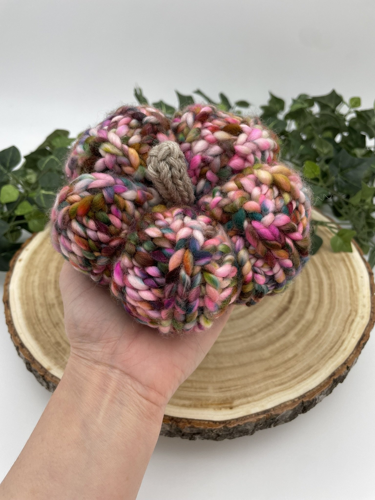 Hand holding a hand-knit pumpkin in fall colors for size reference, showing the colorful yarn and beige stem.