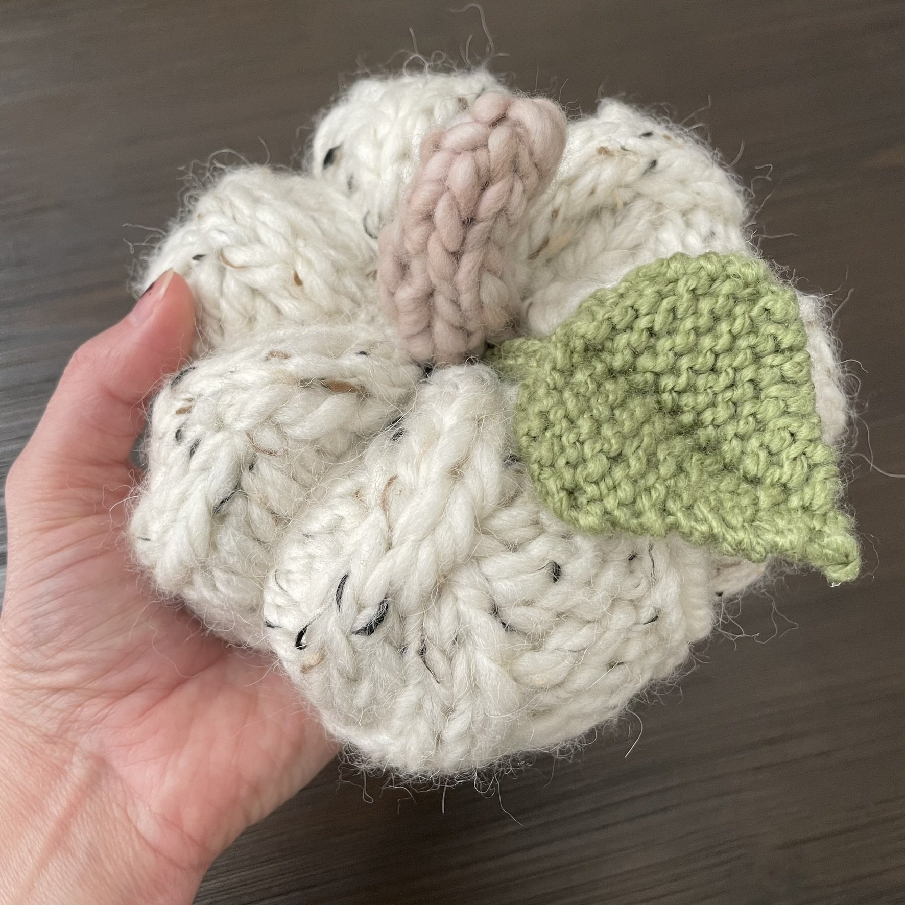 Hand-knit white cable pumpkin placed on a wood base with green leaf and beige stem.